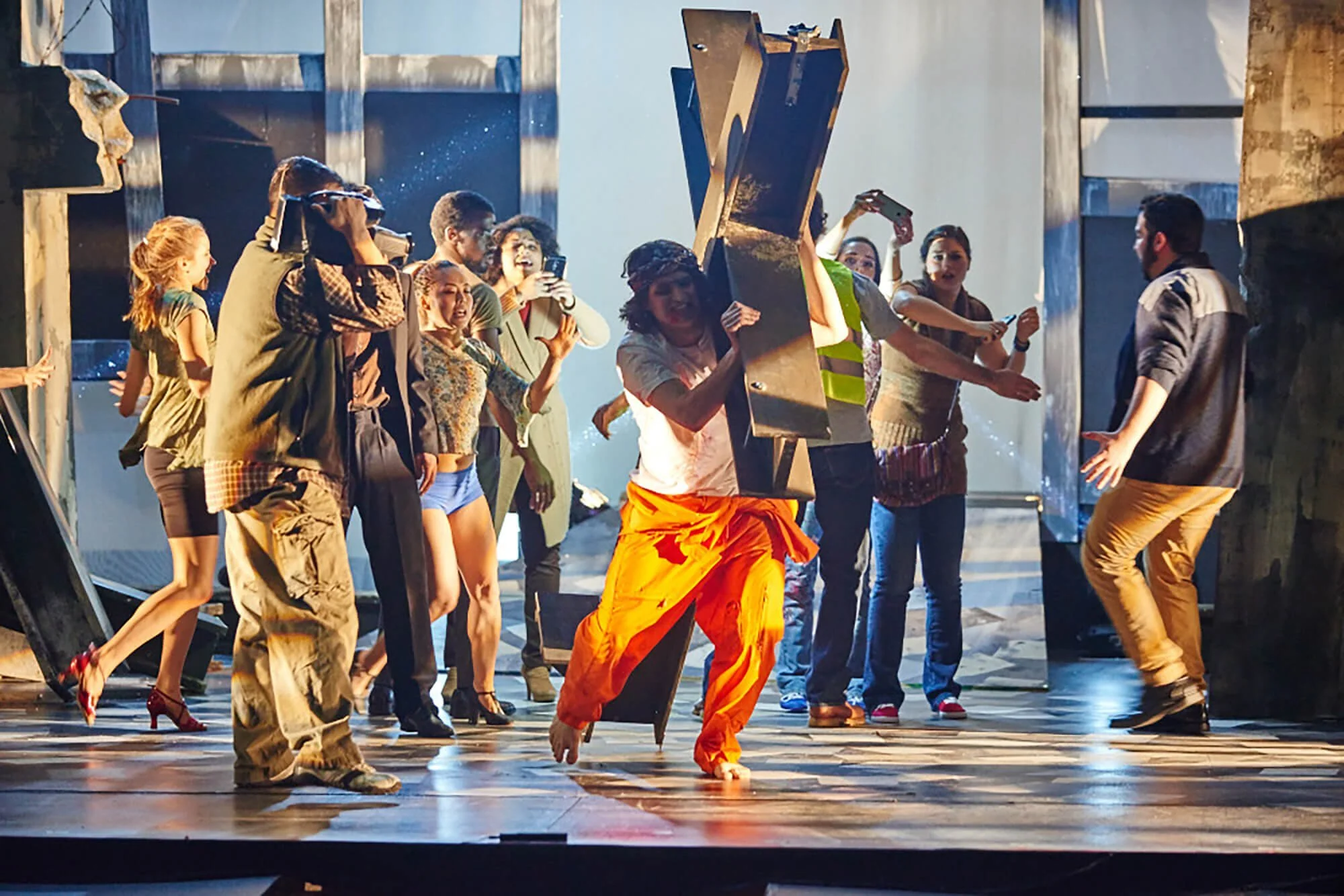 Group of people on stage during a rehearsed performance, some are dancing and others are taking photos or videos, with a woman in bright orange pants at the center.