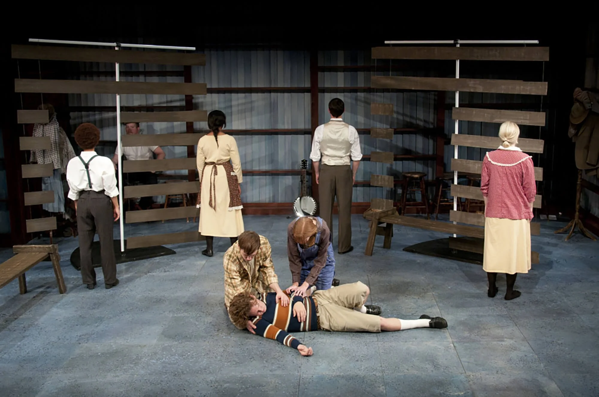 Actors on stage in a theatrical play, some standing and some on the floor, with a set resembling a rustic, old-fashioned environment.