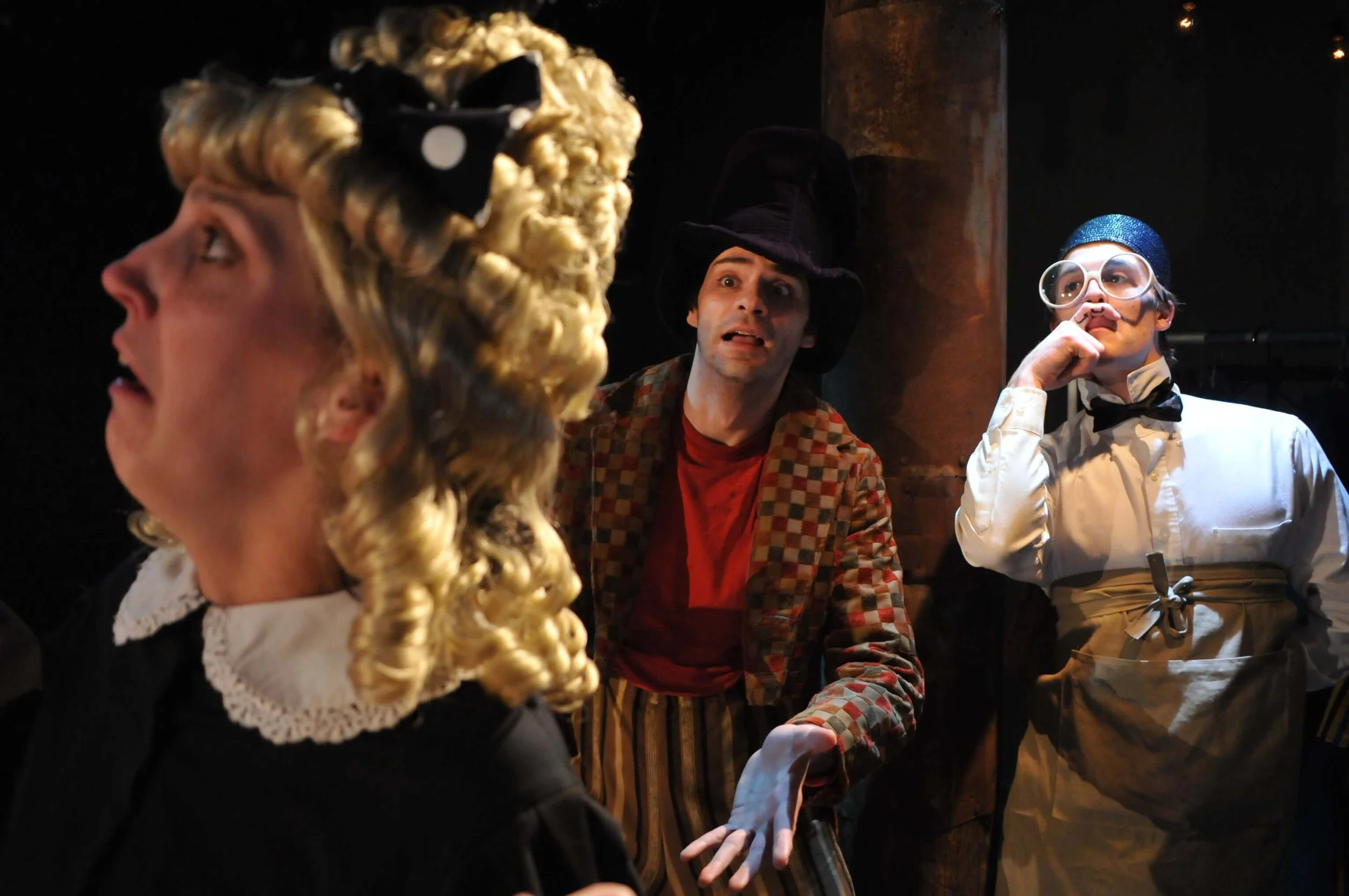 Three actors in costume appear to be performing on stage. The woman on the left has curly blonde hair with a black ribbon and wears a black dress with a white lace collar. The man in the middle wears a tall black hat, a colorful checkered jacket, and