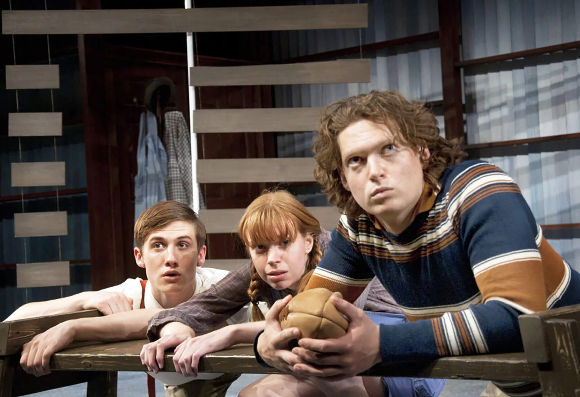 Three young actors, two males and one female, laying on a wooden bench looking intently at something off-camera, with a person holding a ball in the foreground in a room with wooden walls and hanging clothes in the background.