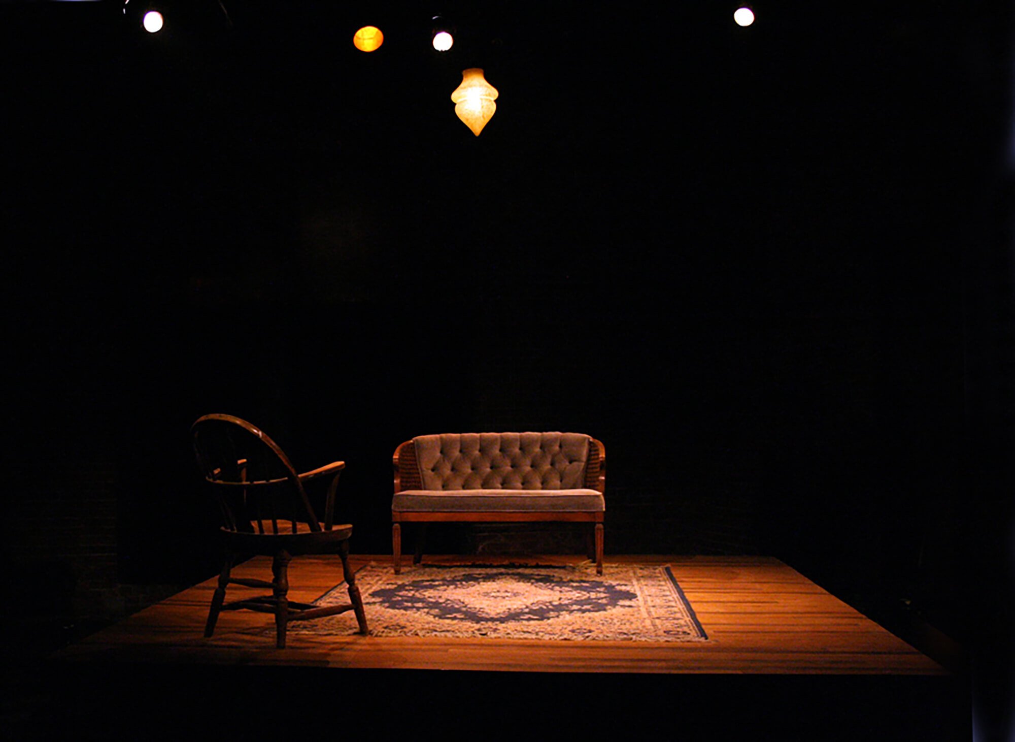 A dimly lit stage with a vintage rug, a wooden armchair, a plush upholstered loveseat, and a hanging pendant light.