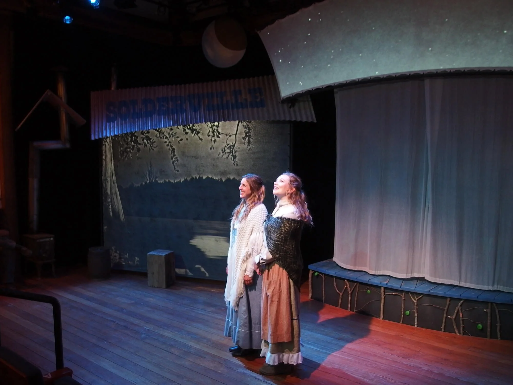 Two women in period costumes stand on a wooden stage, smiling and looking upwards, with a scenic backdrop of trees and water.
