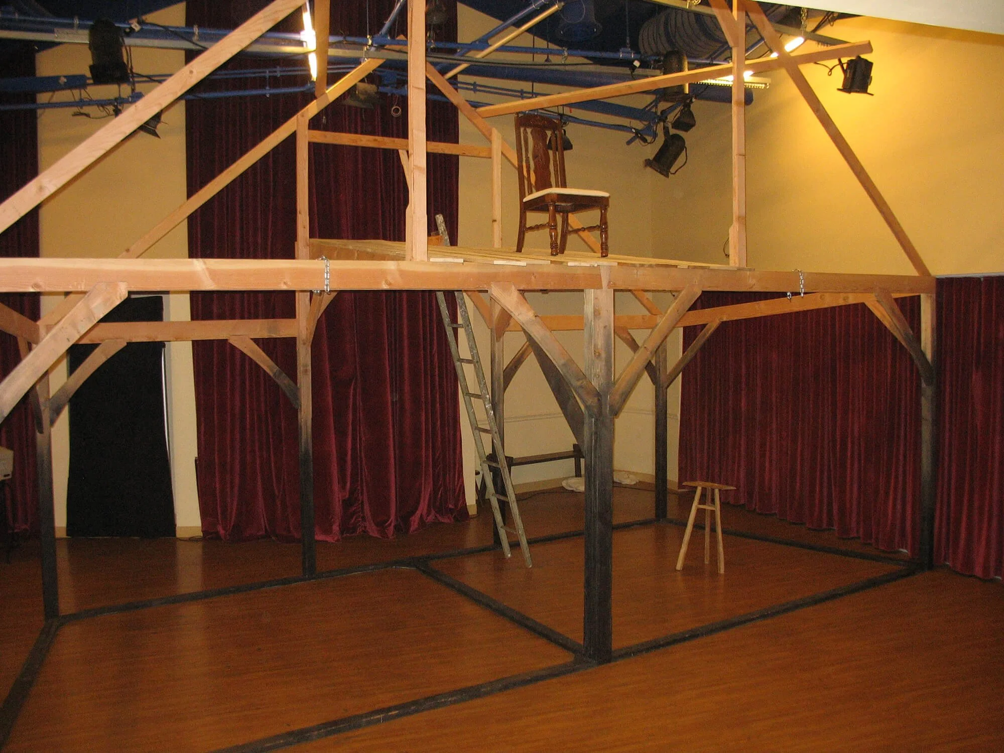 A wooden stage or set construction inside a room with red curtains, a small stool, a ladder, and a chair on top of the stage. Stage lighting is visible overhead.