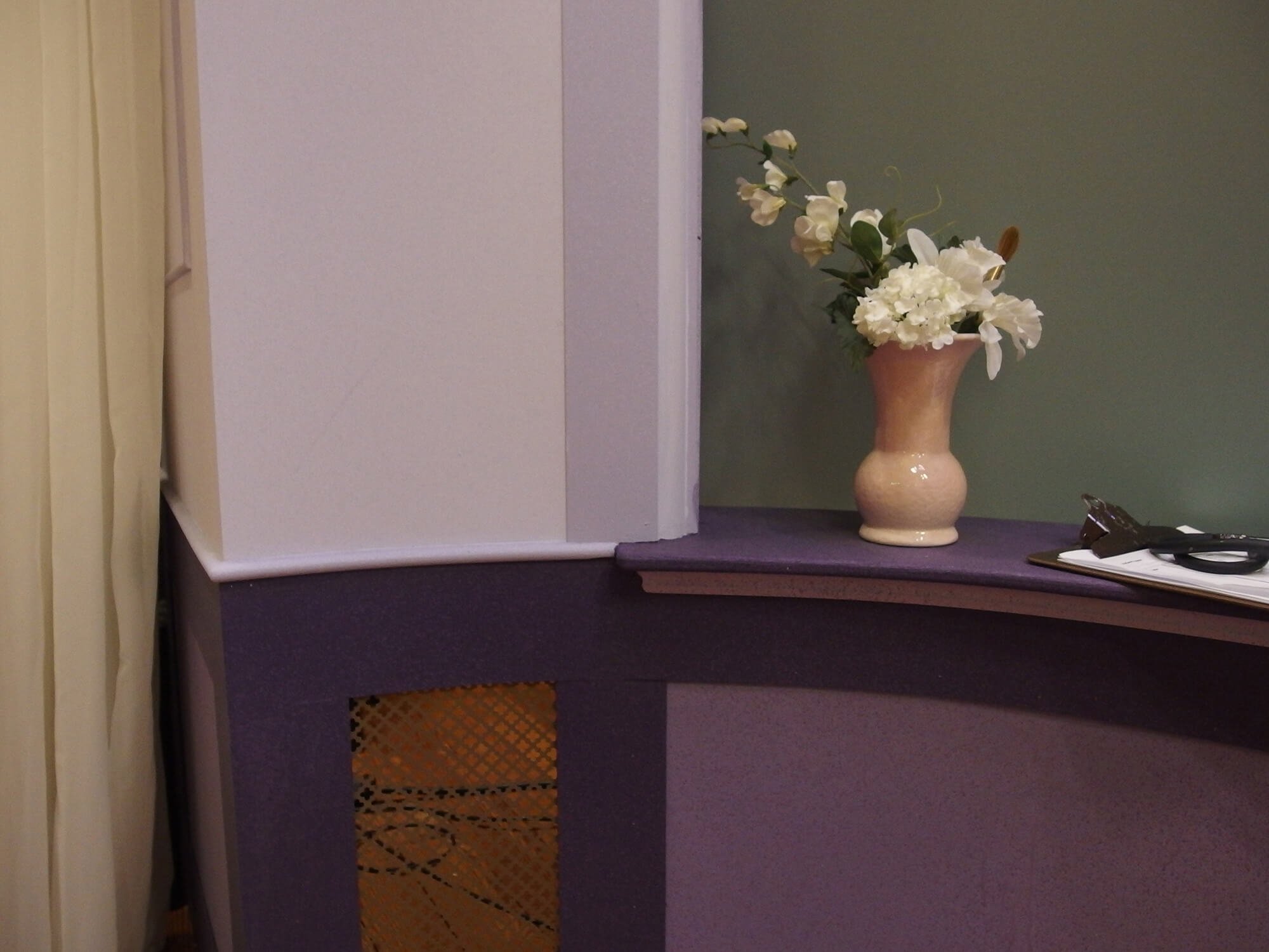 A beige ceramic vase with white flowers and green leaves sitting on a purple countertop with a green wall in the background.