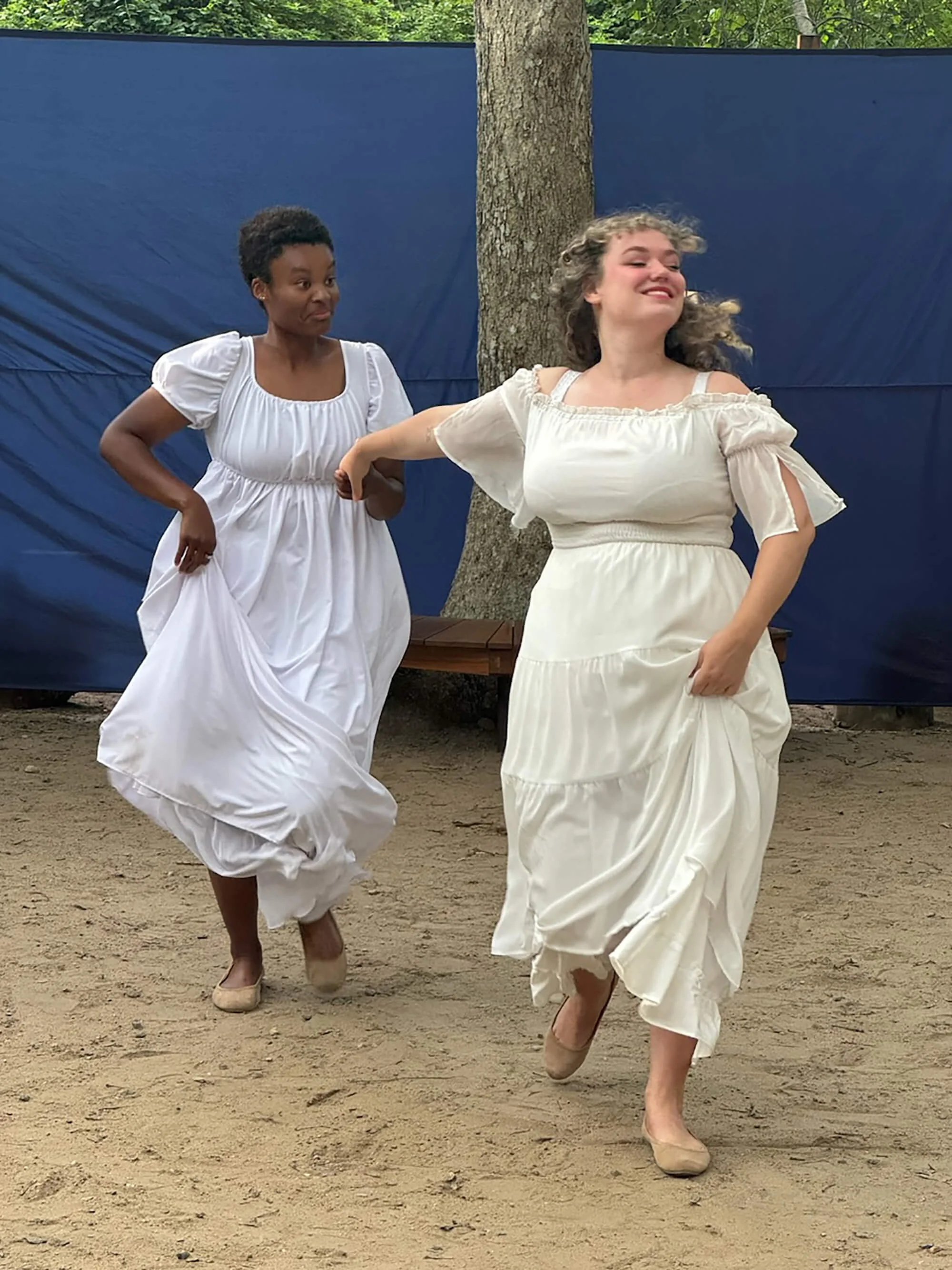 Two women wearing white dresses are dancing outdoors on a dirt ground, with a blue fabric backdrop and trees in the background.