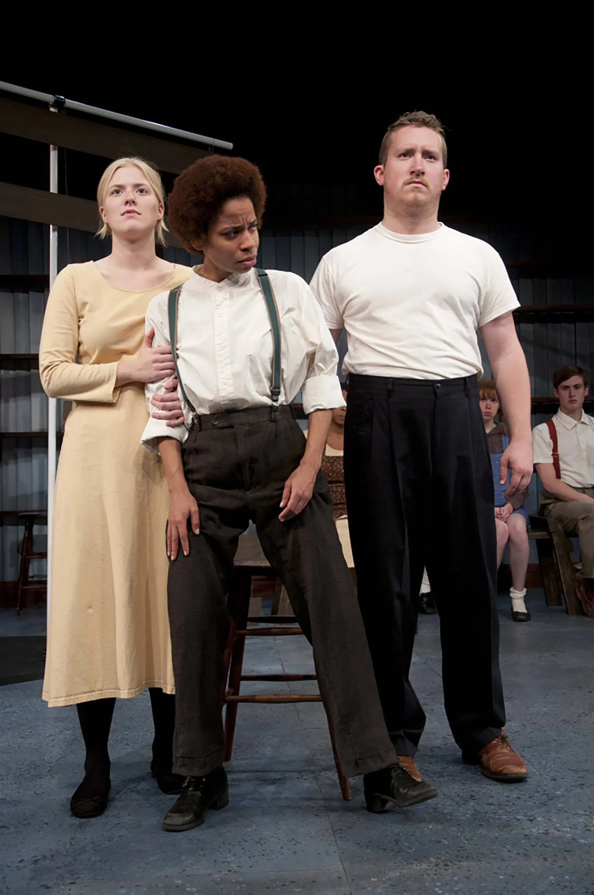 Three actors standing on stage in a theatrical production, with a woman holding onto a woman dressed in vintage clothing at center, and a man standing beside her, all looking serious. Several other actors seated in the background.