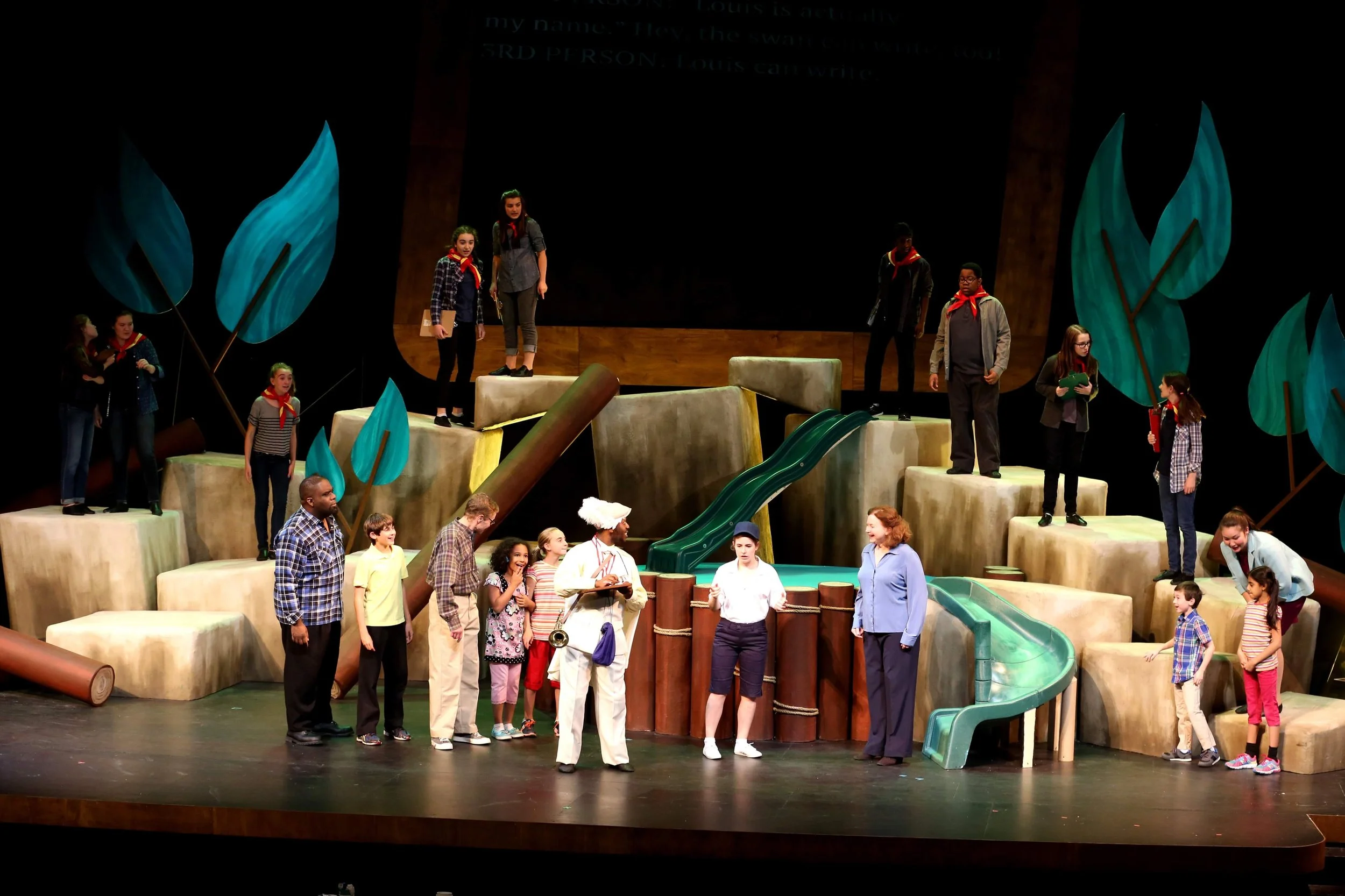 Children performing a play on a stage with large stylized trees and maze-like structures, some children standing on elevated platforms and others on the stage level, with adults interacting with the children.