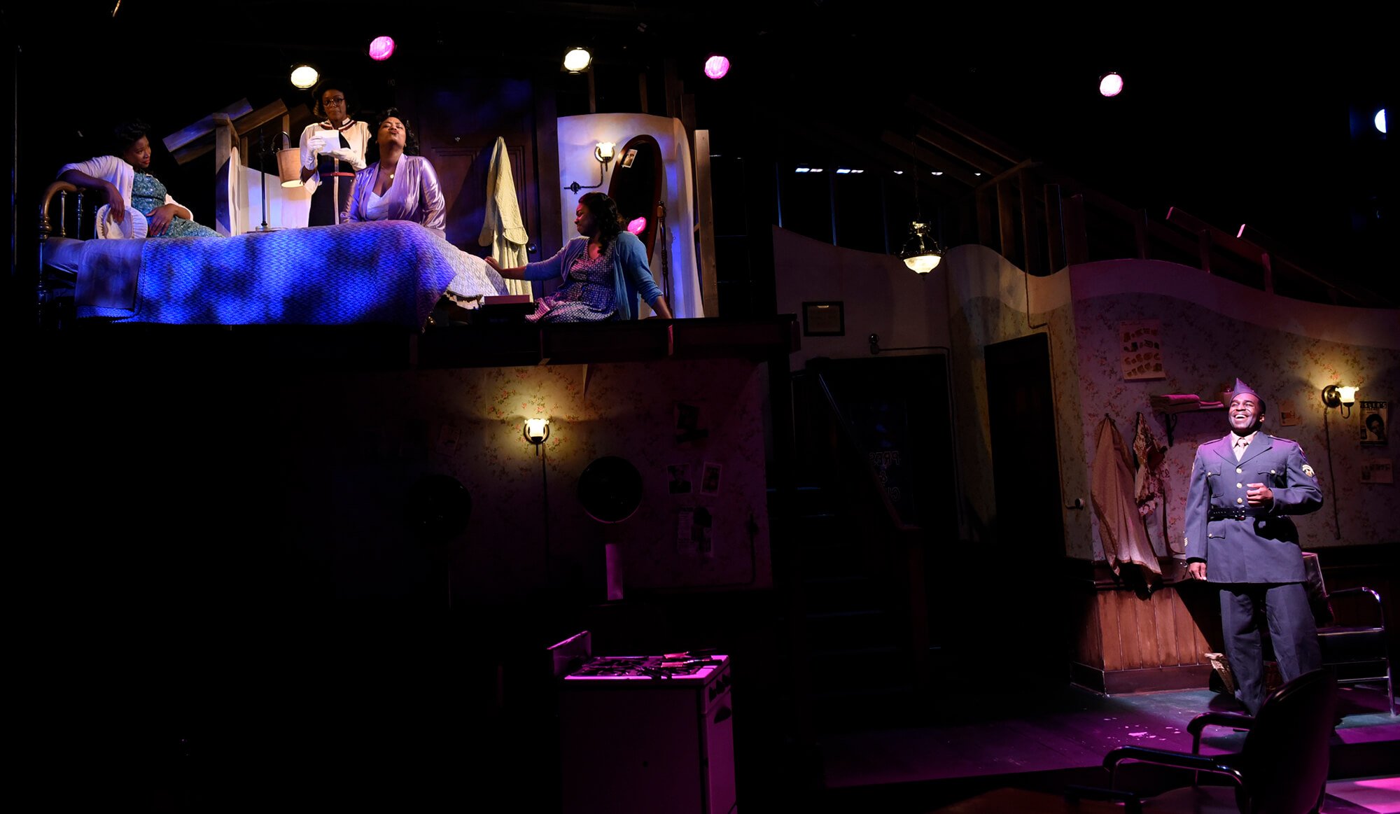 The stage depicts a scene from a theatrical play with four women on an elevated area, engaged in a discussion, and a man in a police uniform standing below, smiling. The setting resembles a bedroom with furniture and decor, illuminated by stage light