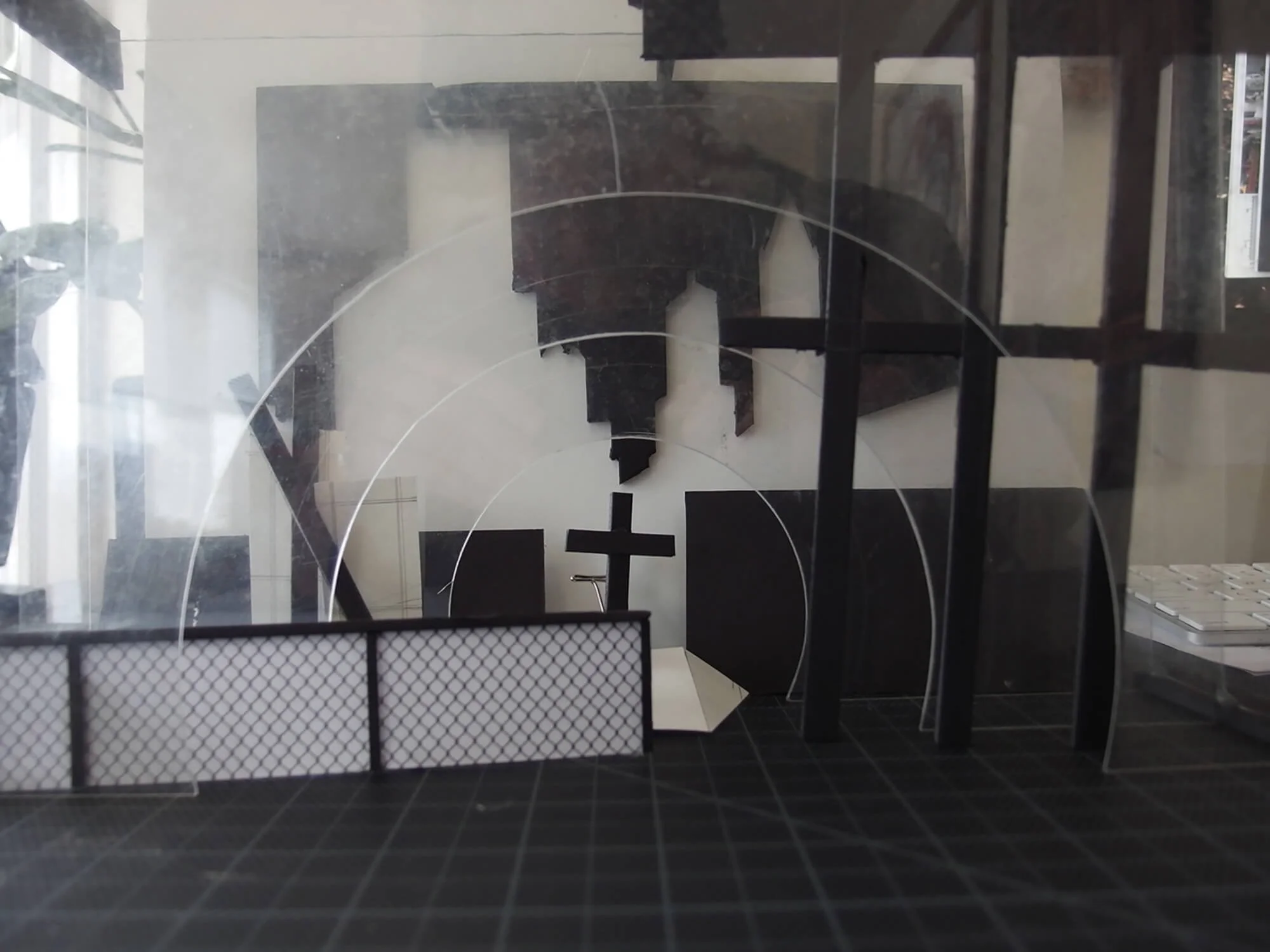 A small model of a church or chapel with a cross at the front, black roof, and white walls, viewed through a glass surface with lines and reflections, inside a room with various objects, including a scale model and a keyboard.