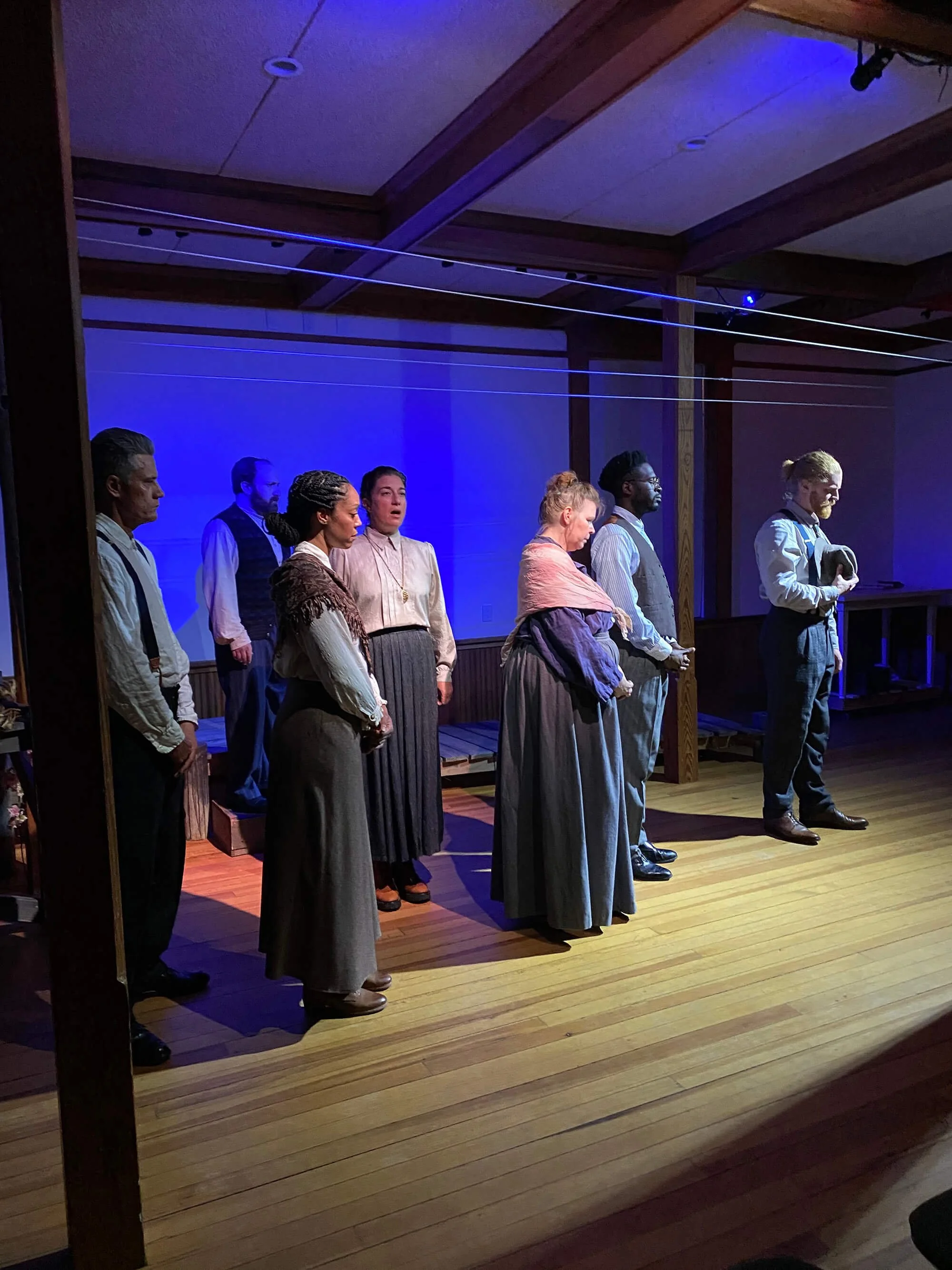 A scene from a theatrical play featuring eight actors dressed in historical costume, standing on a wooden stage with blue and purple lighting. The actors are arranged in a line, with some facing forward and others looking sideways.