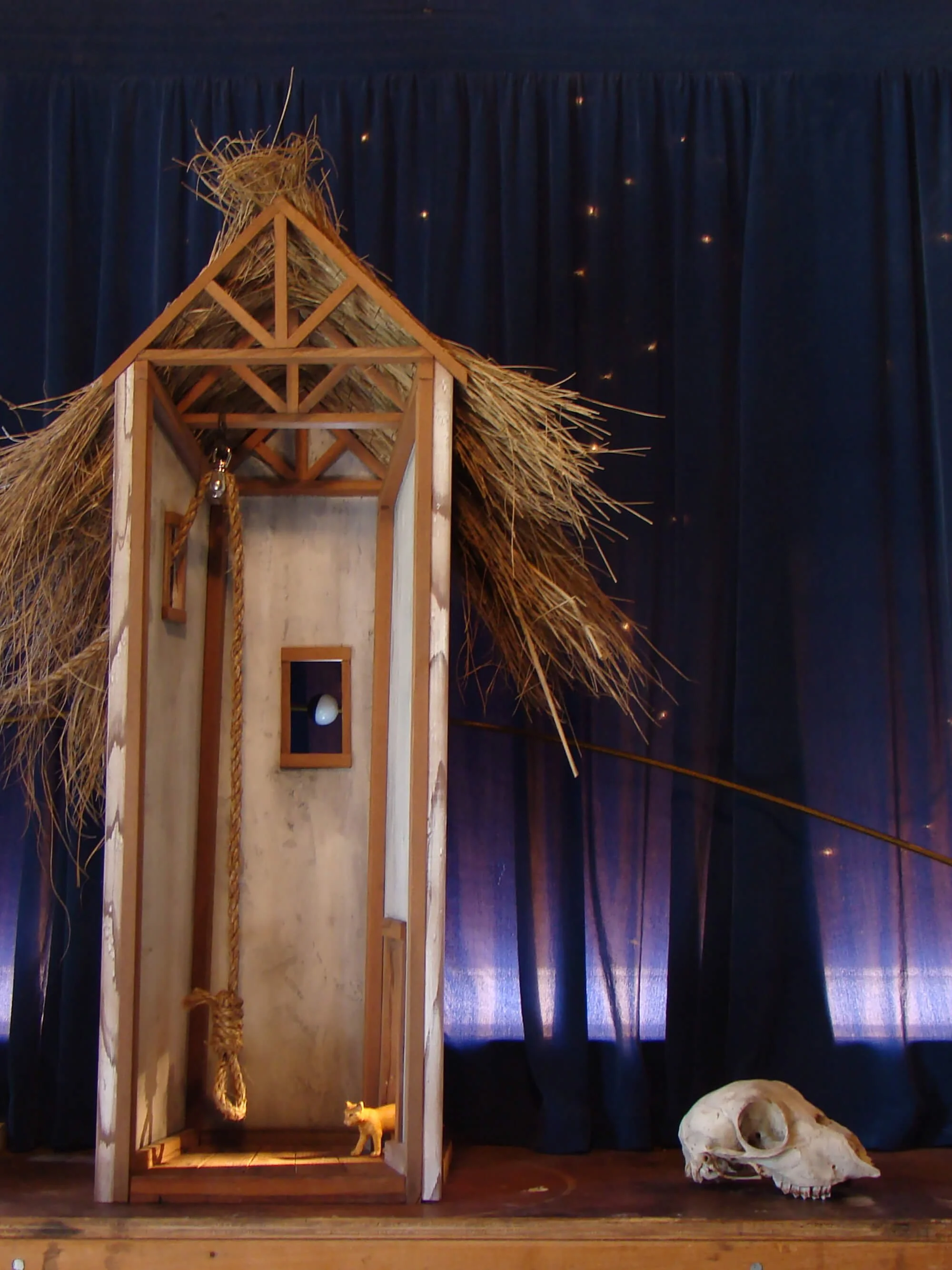 A small, handcrafted model of a barn with a straw roof, a tiny animal figurine inside, a skull on the ground beside it, and a dark curtain with small lights in the background.
