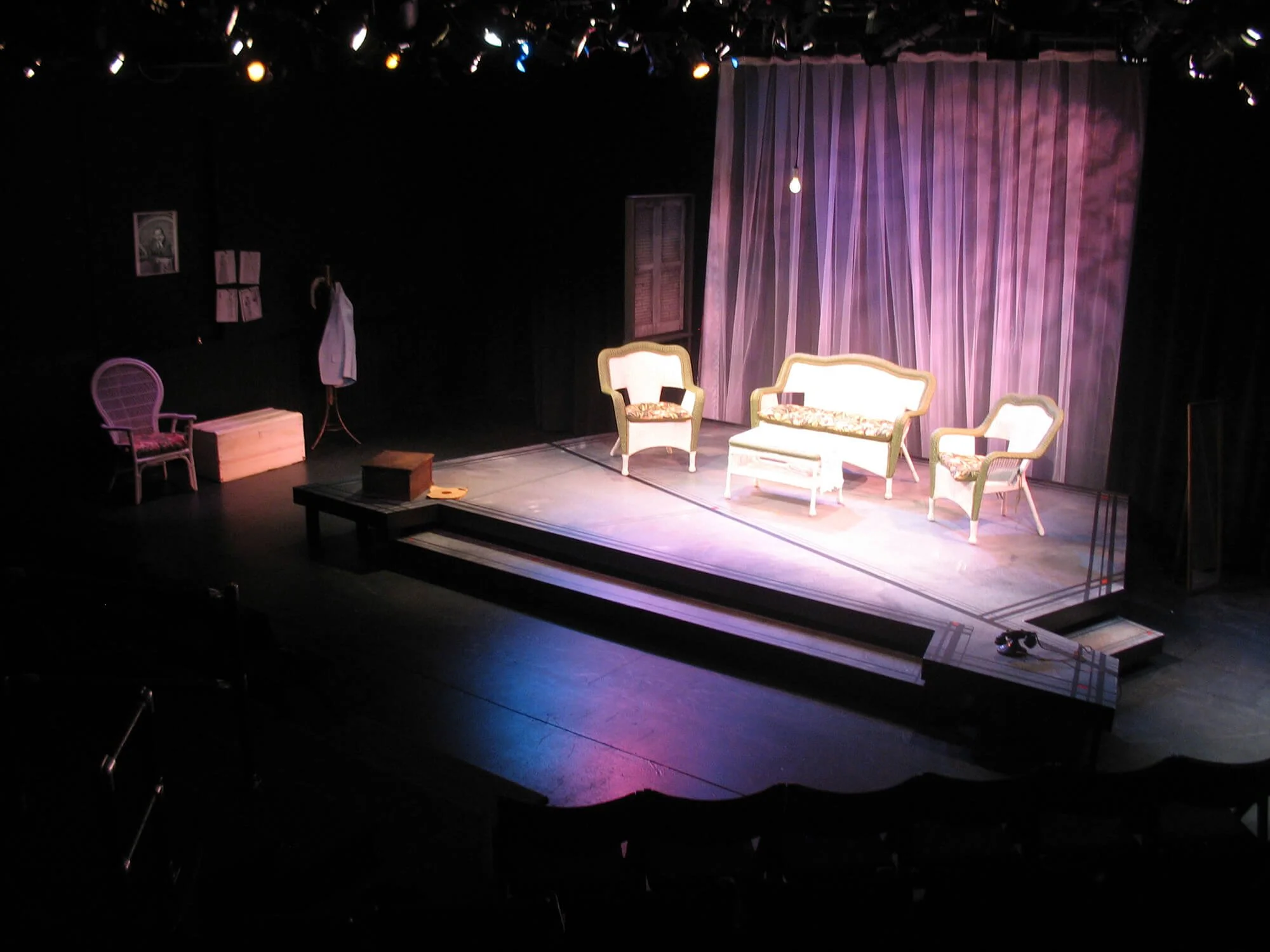 Stage set-up for a play with vintage furniture including a sofa, armchairs, a bench, and a small table with a box. The backdrop is a delicate pink curtain. Stage lighting highlights the furniture while other parts are dimly lit.