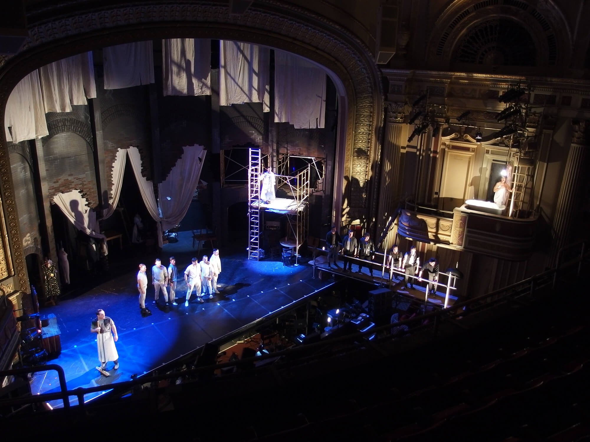 Theater stage with performers and crew, set in an ornate auditorium with gold accents, stage scaffolding, and hanging curtains.