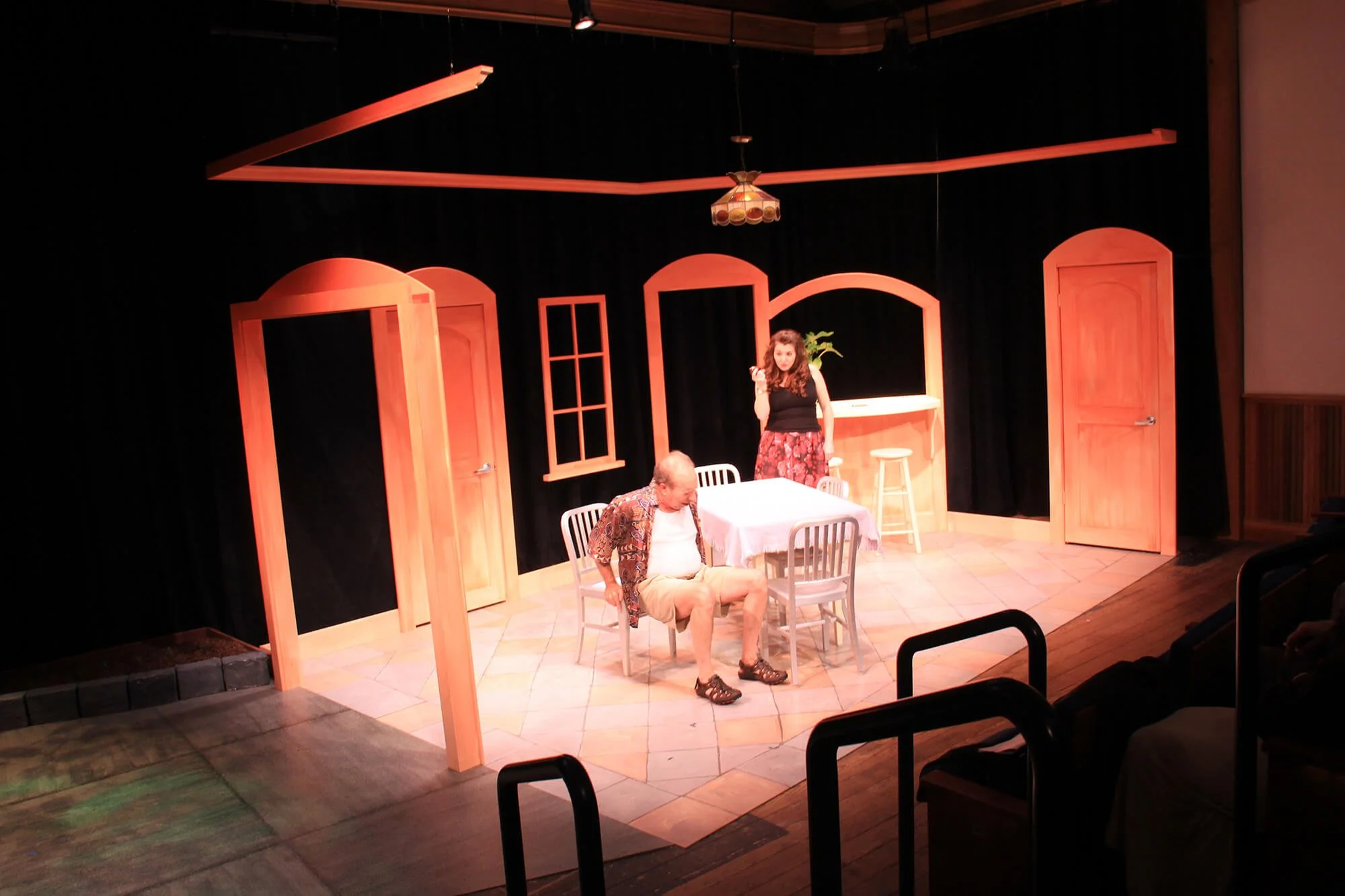 A theatrical stage with two actors, one sitting at a table and the other standing nearby, set in a room with wooden doors and windows, a small bar or counter, and a black curtain backdrop.