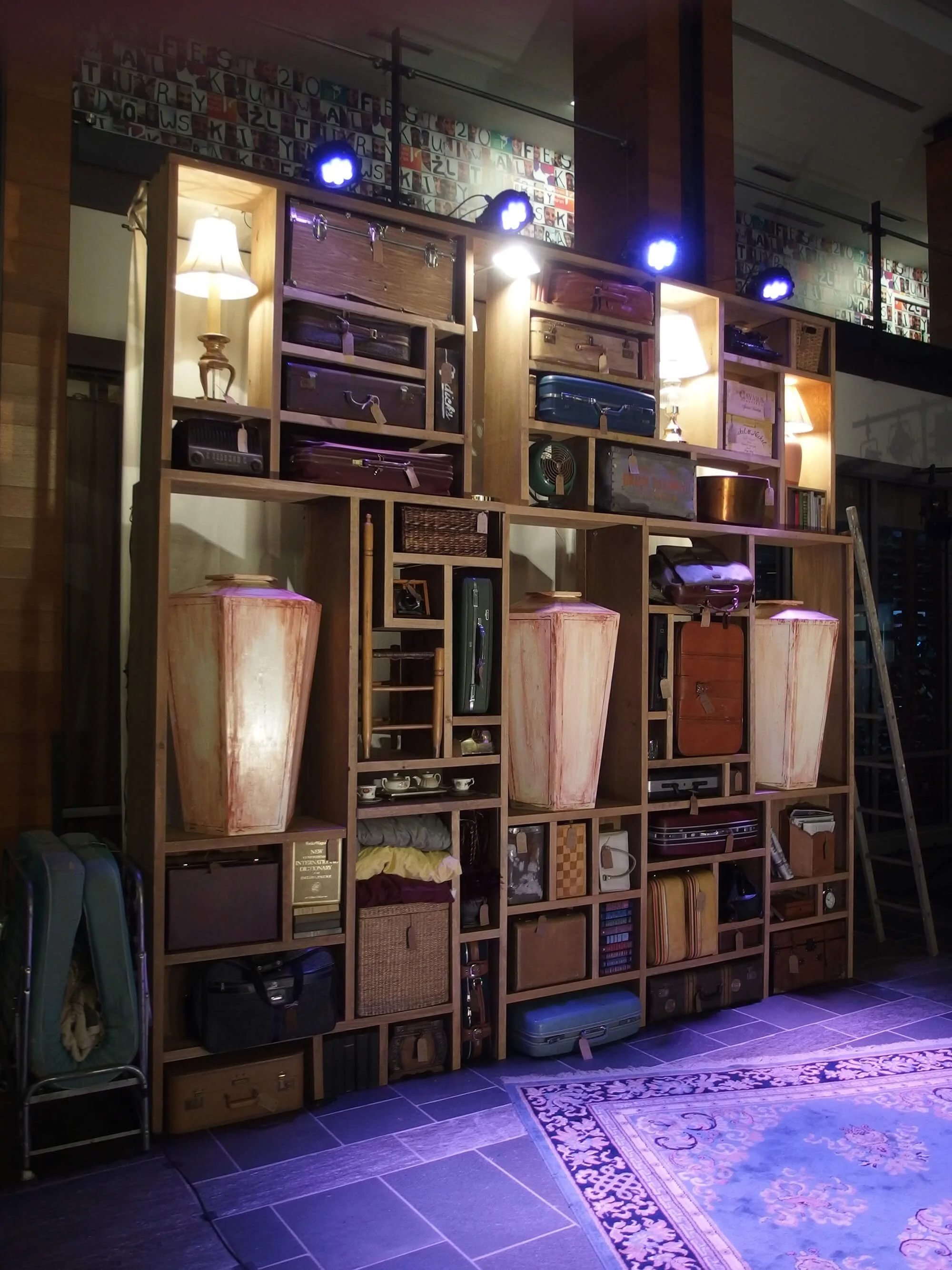 A large wooden shelving unit filled with vintage suitcases, lamps, books, and decorative items. Two lamps are lit, illuminating the shelves. There are purple and blue lights above. A rug is partially visible on the dark tiled floor in front of the sh