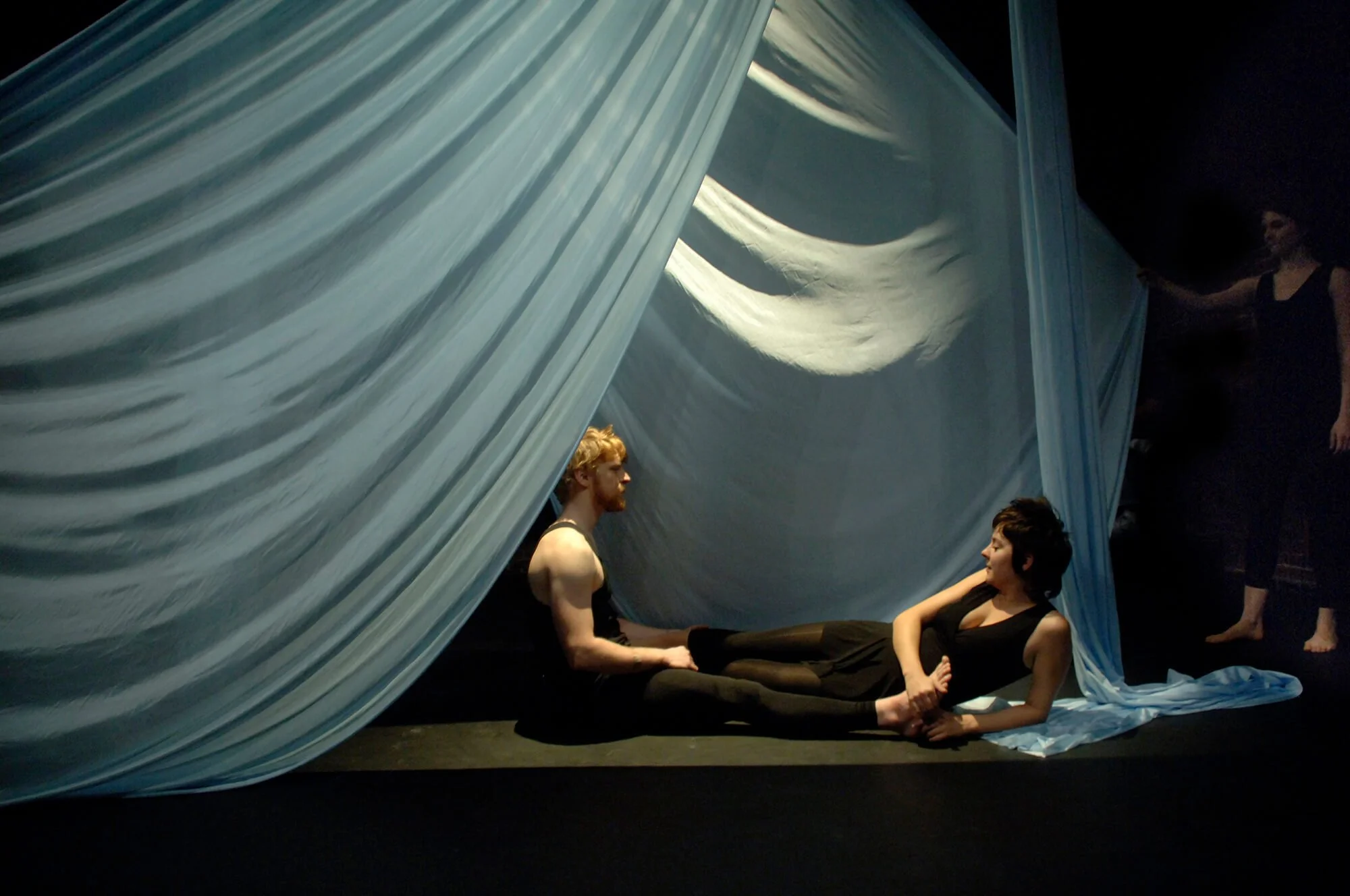 Two actors on stage, one man sitting cross-legged and a woman reclining on her side with her head propped on her hand, under a large flowing white curtain with a moon-shaped opening.