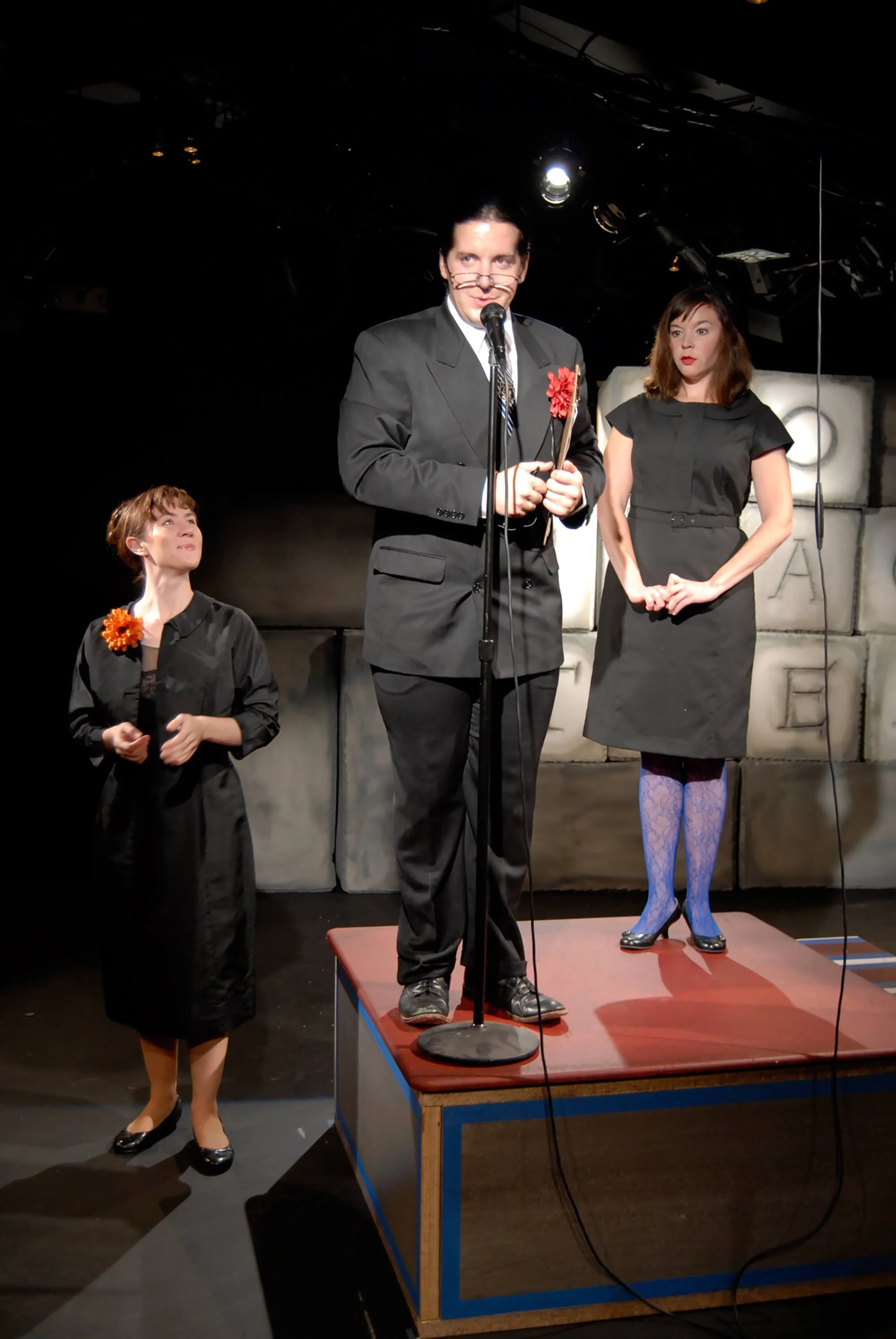 Three actors on a stage during a theatrical performance, with one man standing on a raised platform speaking into a microphone and two women standing nearby, one on the ground and the other on the platform, with a backdrop of gray bricks.