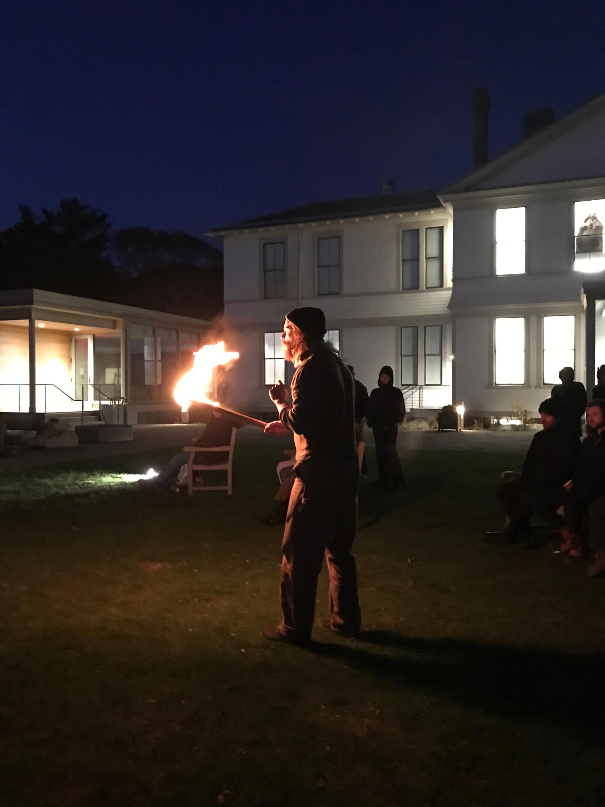 Person performing fire-breathing during an outdoor evening gathering, with a lit torch in hand and a house in the background.