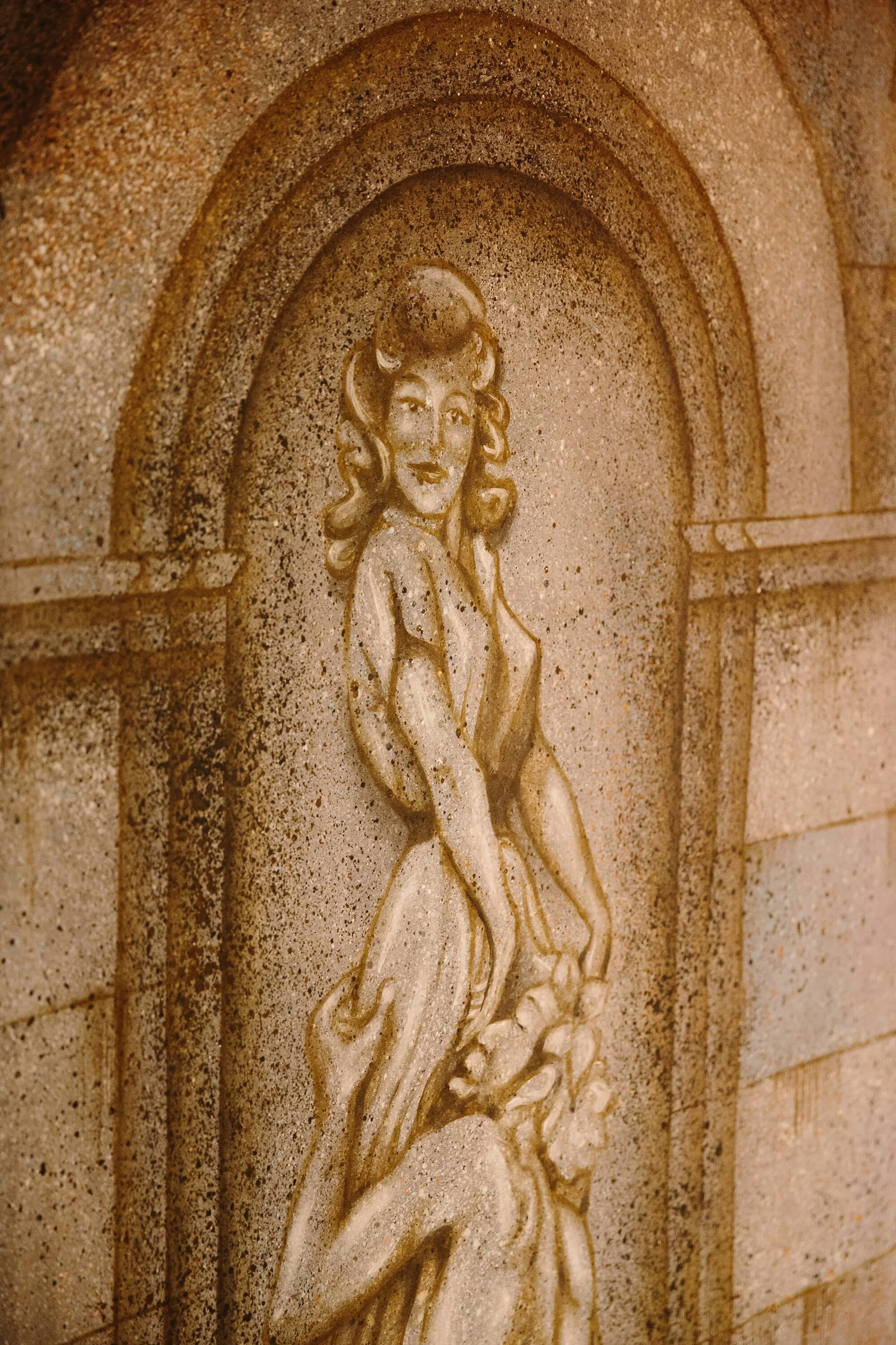 A carved stone relief of a woman with long hair, smiling, standing among flowers, set into a decorative arched wall.