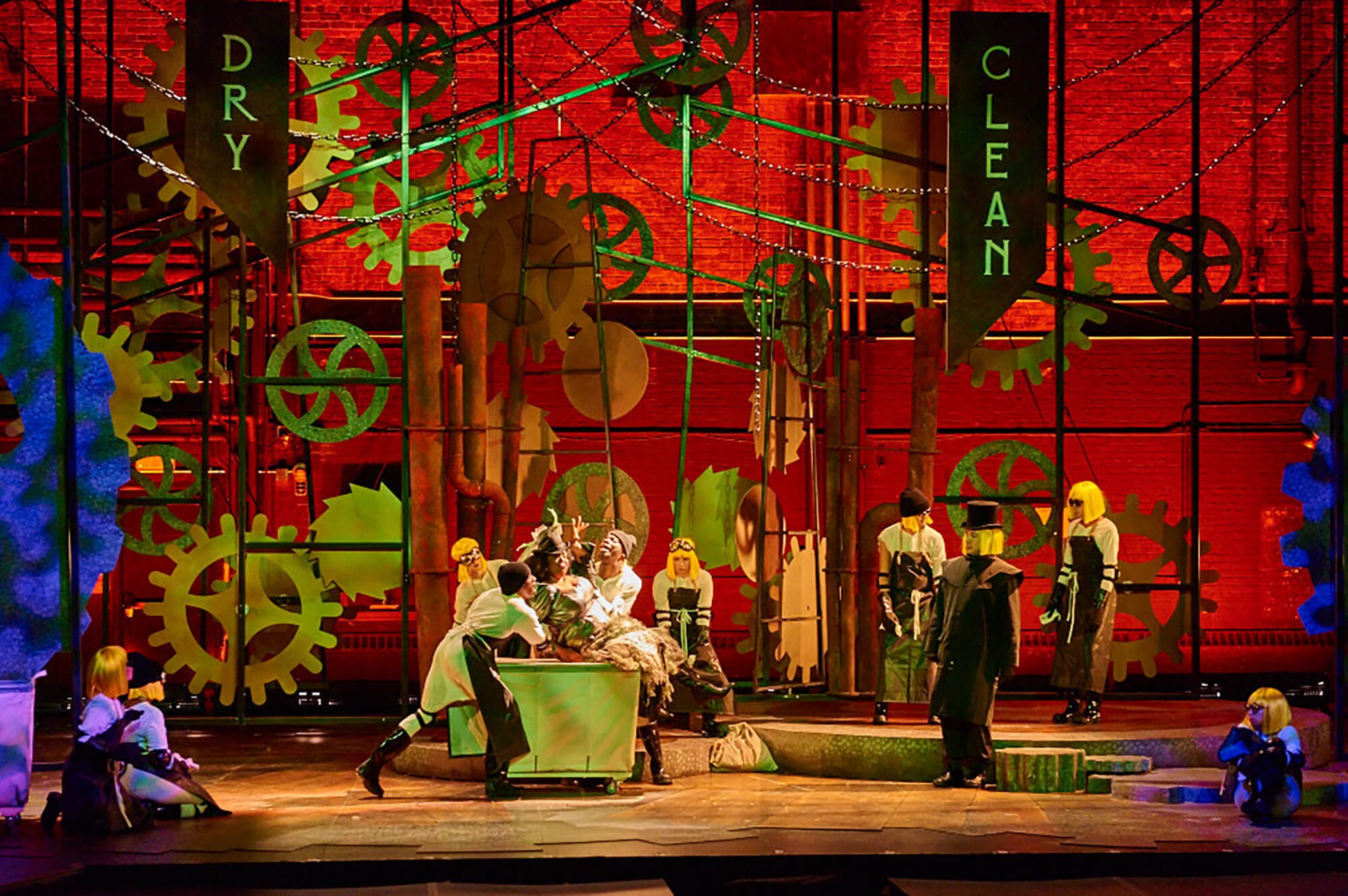 A theatrical stage scene with performers in costumes in front of a backdrop of oversized gears and cogs, with signs reading 'Dry' and 'Clean.'