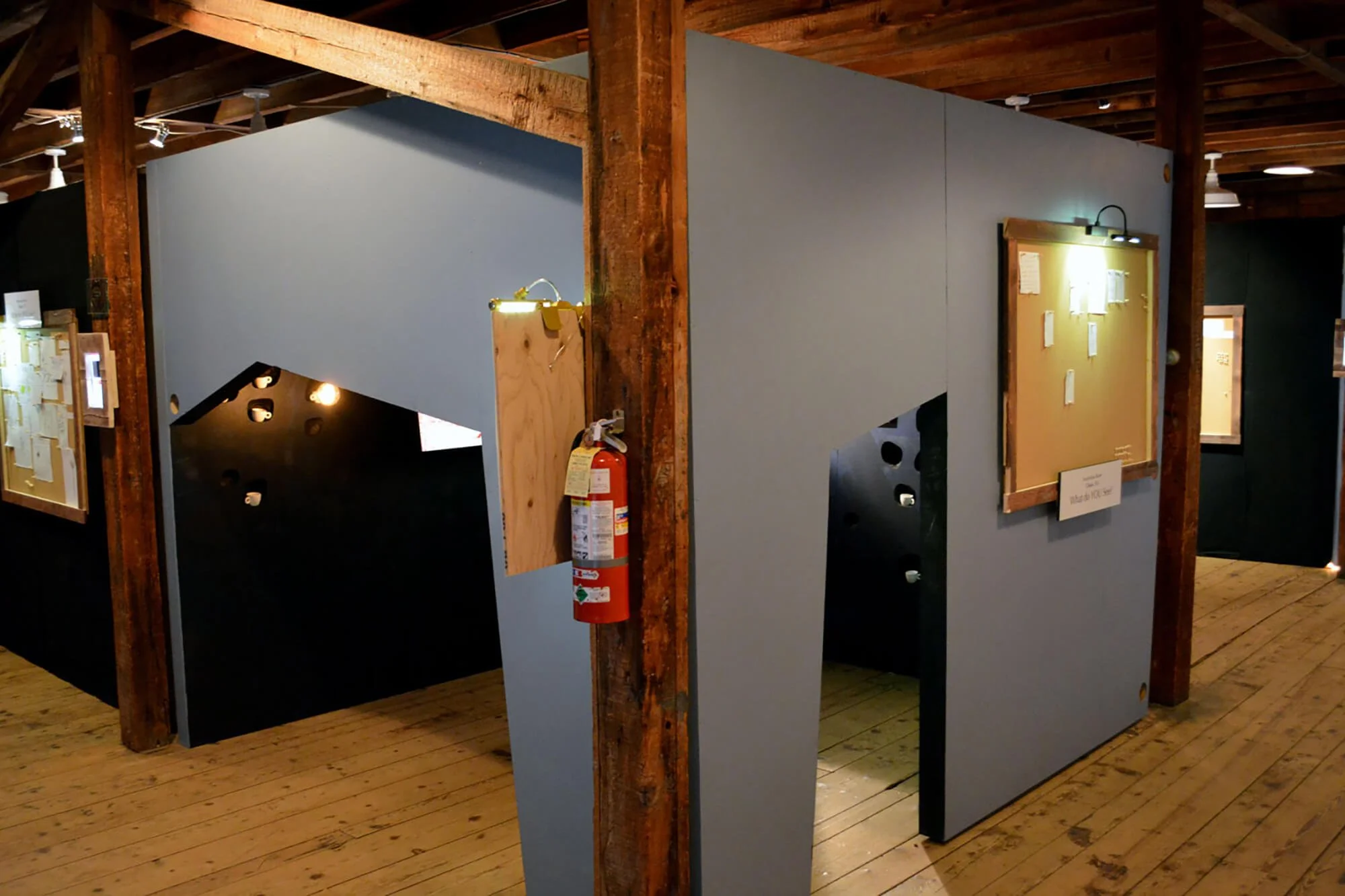 Interior of an art exhibit with wooden beams, display boards, and a fire extinguisher attached to a corner pillar.