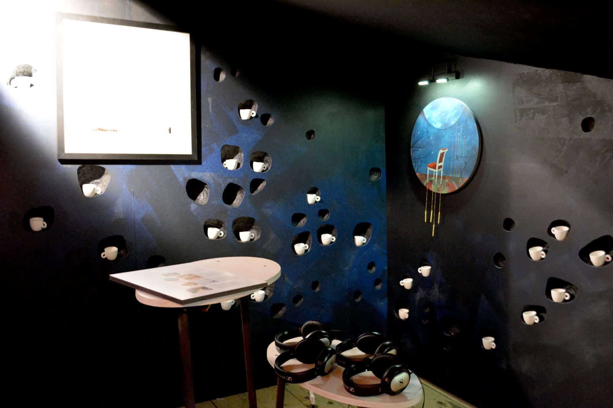A dark room with a wall decorated with many small white coffee cups, some placed in wall cutouts and others hanging. There is a small table with a tablecloth and a set of black headphones on top. A large square bright screen is on the left side, and 