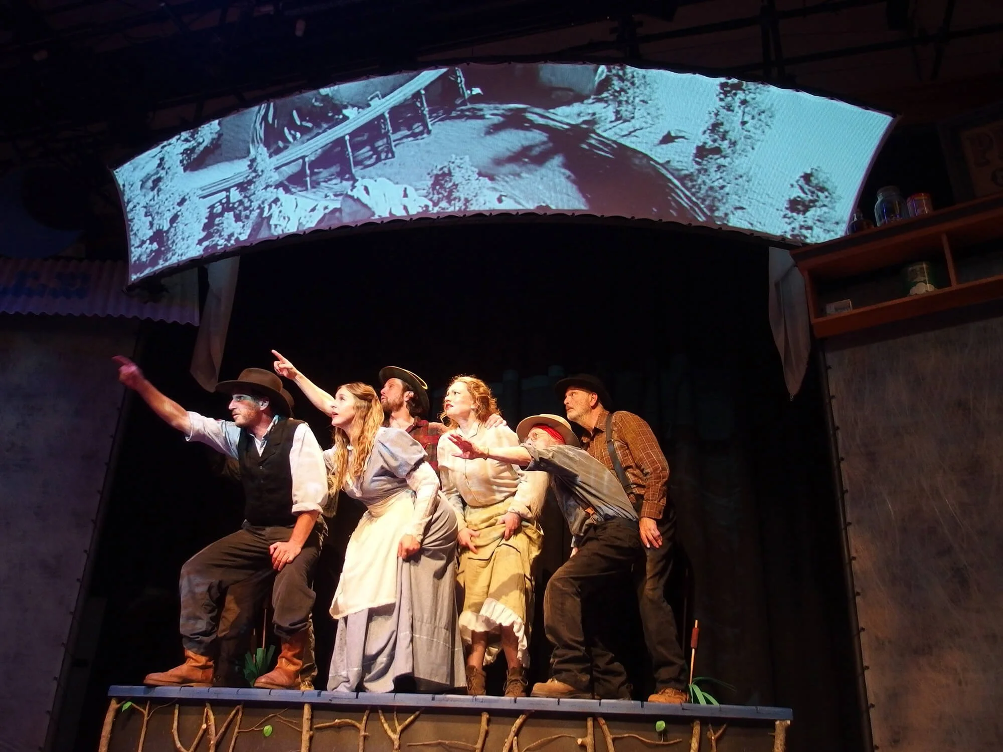 Six actors dressed in period costumes performing on a stage with a black background. They are looking and pointing upward, with a large curved screen above them showing a black and white scene.