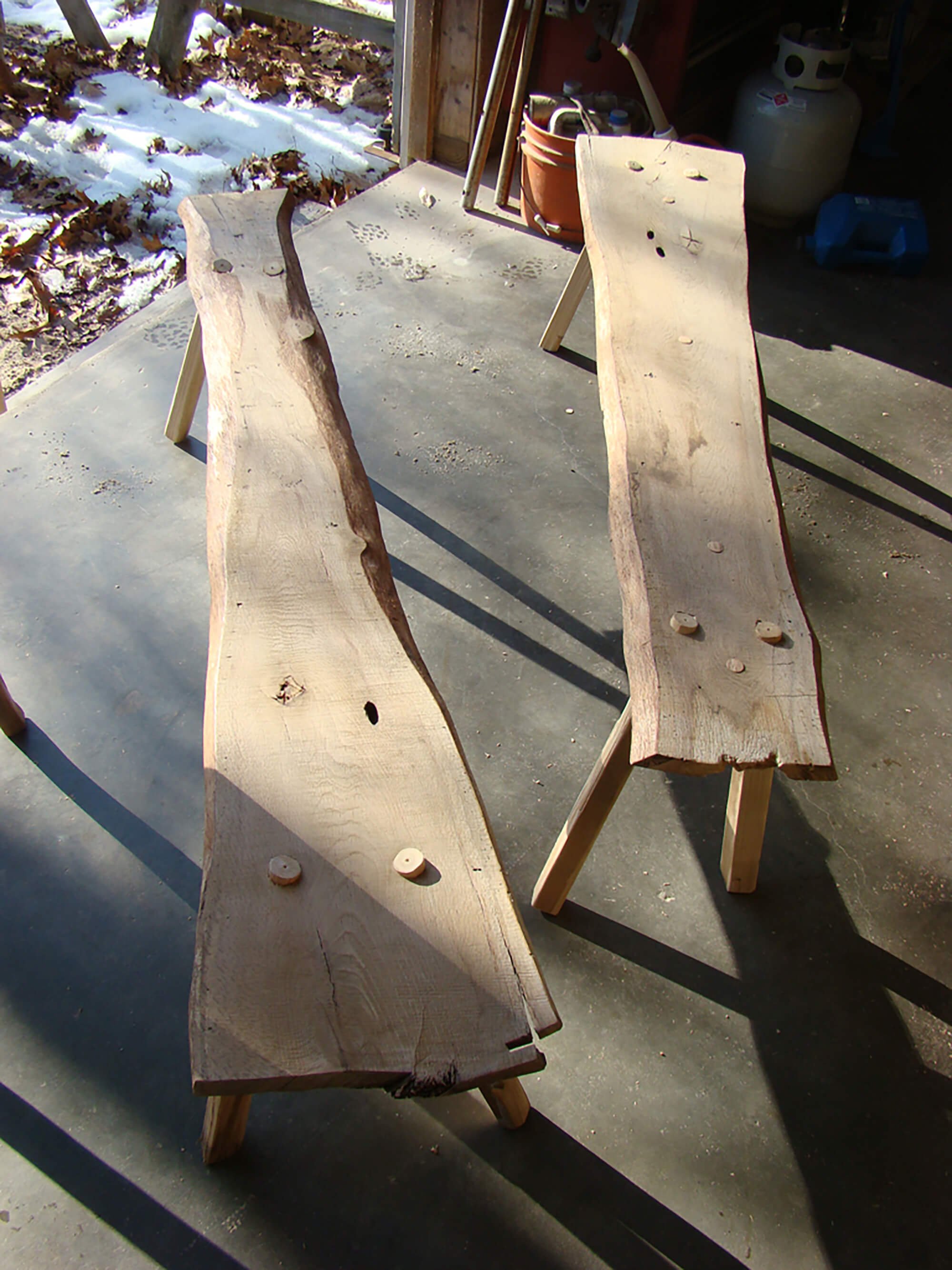 Two wooden benches made from large, natural wood slabs with live edges, supported by simple wooden legs, placed outdoors on a concrete surface with snow and fallen leaves in the background.