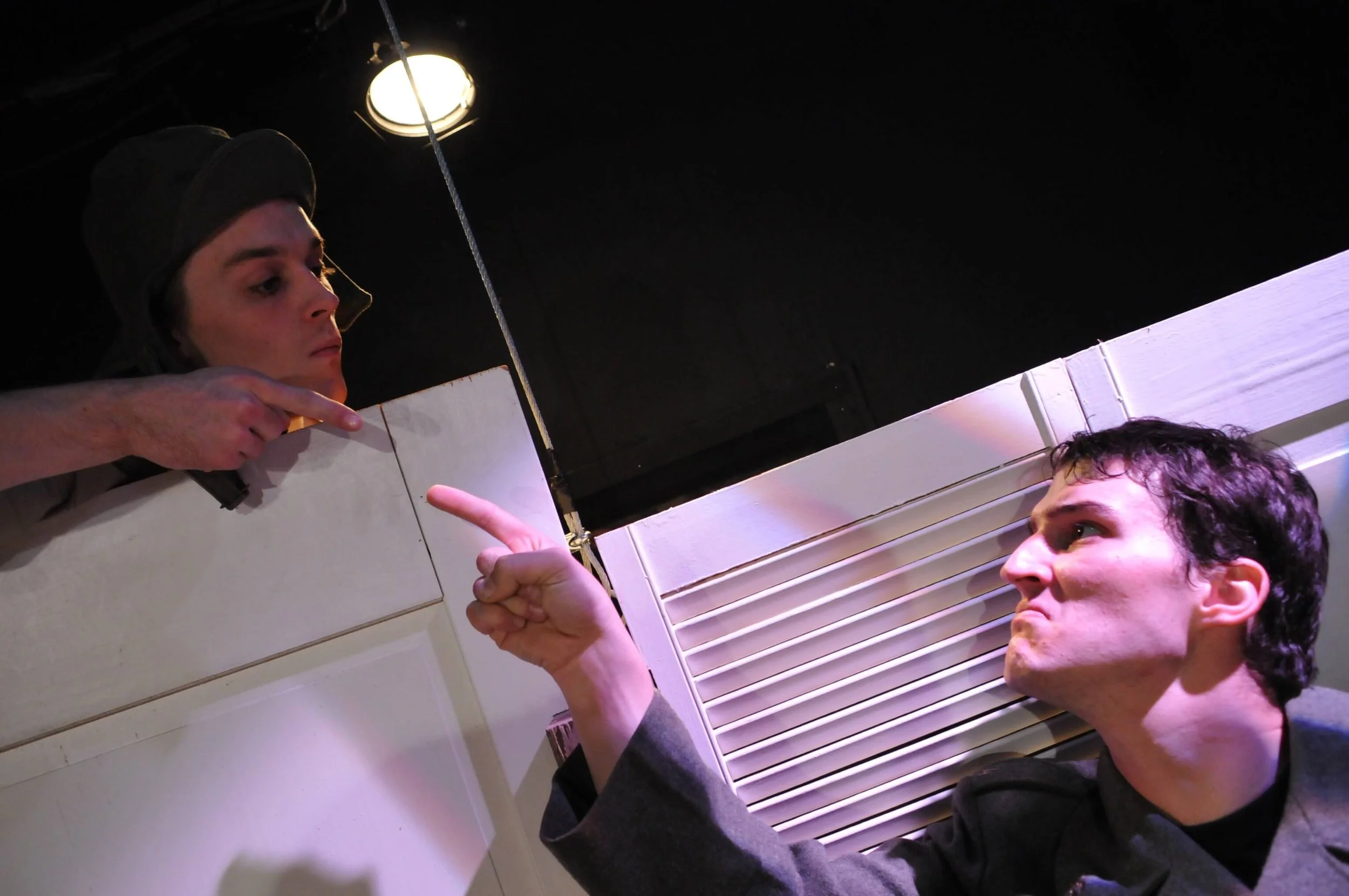 A woman and a man in a tense interaction, with the woman pointing at the man behind a white partition and the man angrily pointing back, in a dramatic scene.