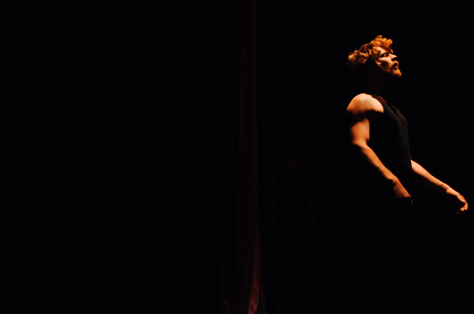 A person with curly hair and a beard, wearing a sleeveless top, is illuminated from the side in a mostly dark environment, creating a dramatic silhouette.
