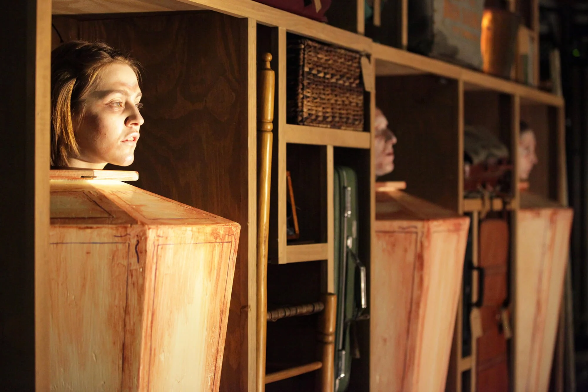 Side view of a woman’s head and shoulders with her face illuminated, peeking out from a hollowed-out wooden box or structure, with a background of shelves filled with various items.