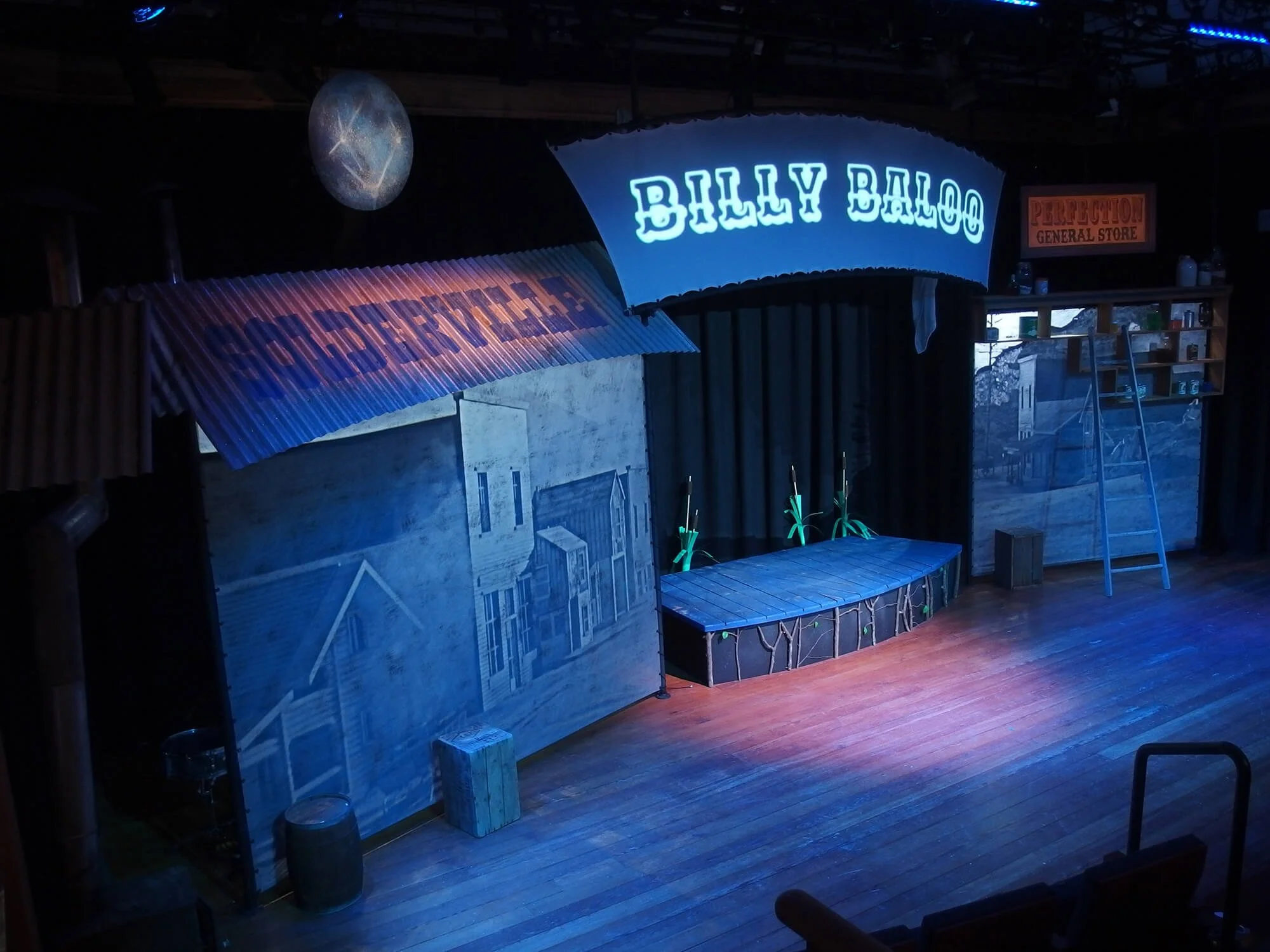 Empty stage with set design for a play, featuring signs reading 'Billy Baloo' and 'Perretion General Store', a backdrop of a building and street scene, a ladder, and props like barrels and crates.