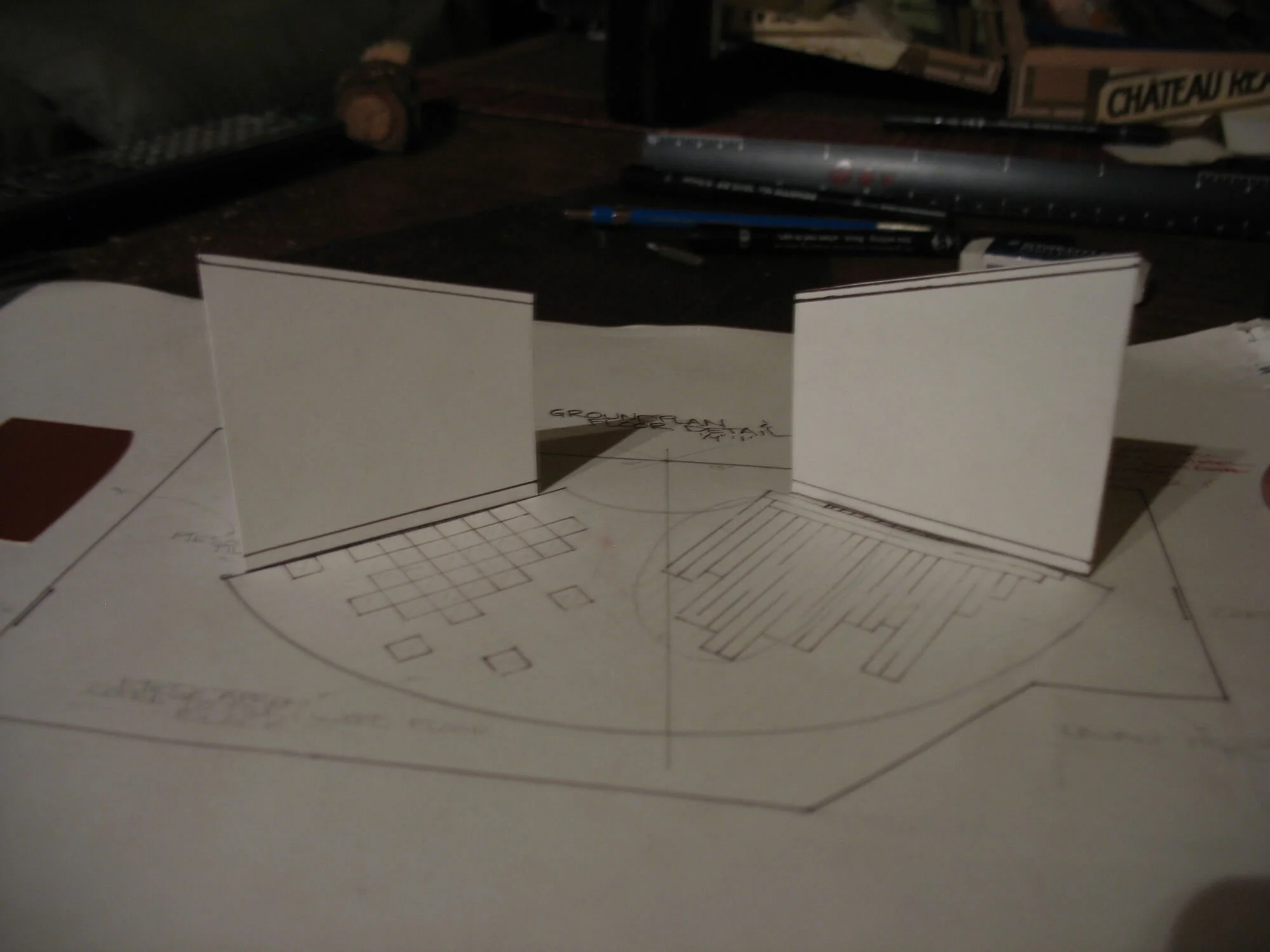 A small architectural model with white paper walls on a hand-drawn floor plan, with various pens and tools in the background.