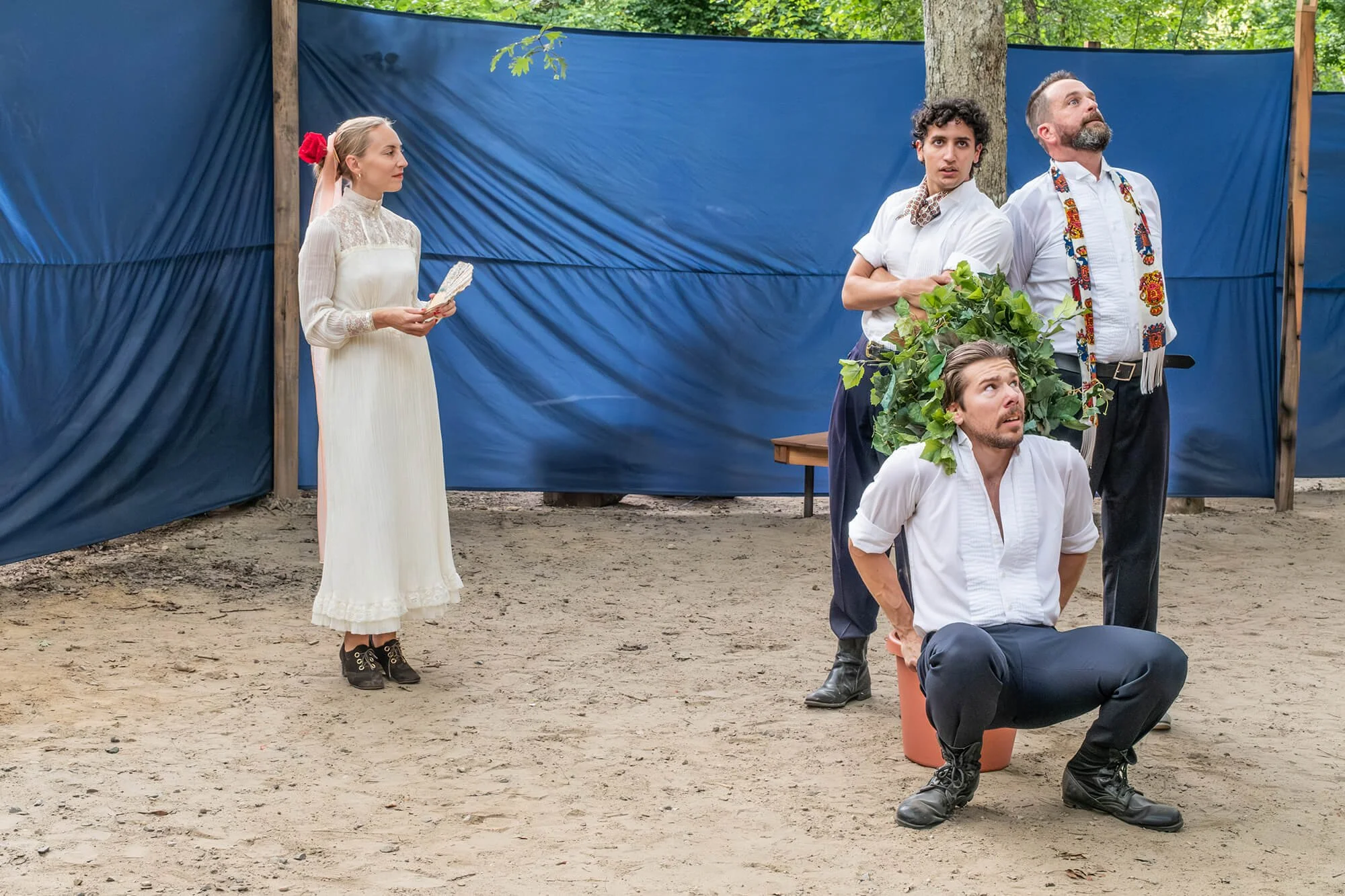 Individuals dressed in theatrical costumes participating in a stage performance outdoors, with a blue fabric backdrop and natural surroundings.