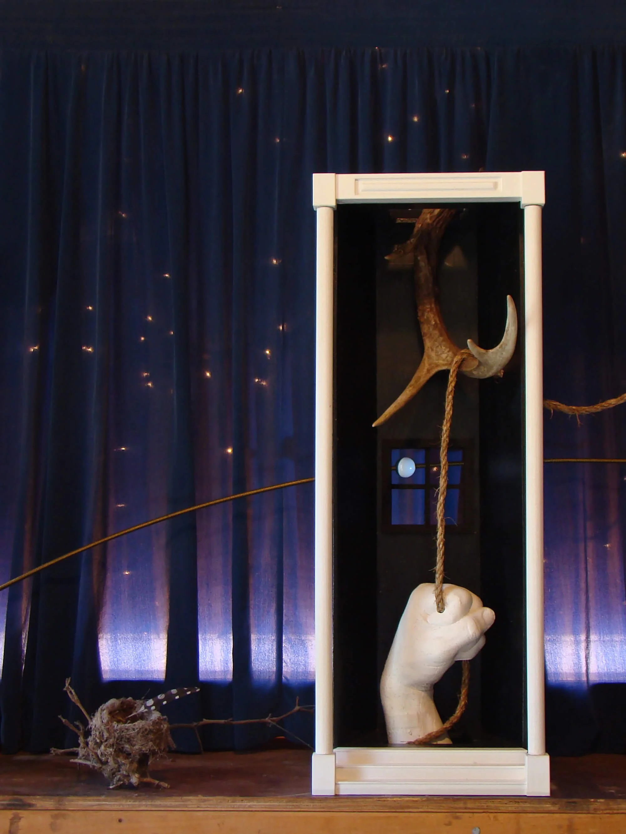 An art installation featuring a large, white, rubbery fist gripping a rope inside a glass case. A crescent moon and a deer antler are also suspended inside the case. The background includes dark blue curtains with small lights, and a small bird's nes