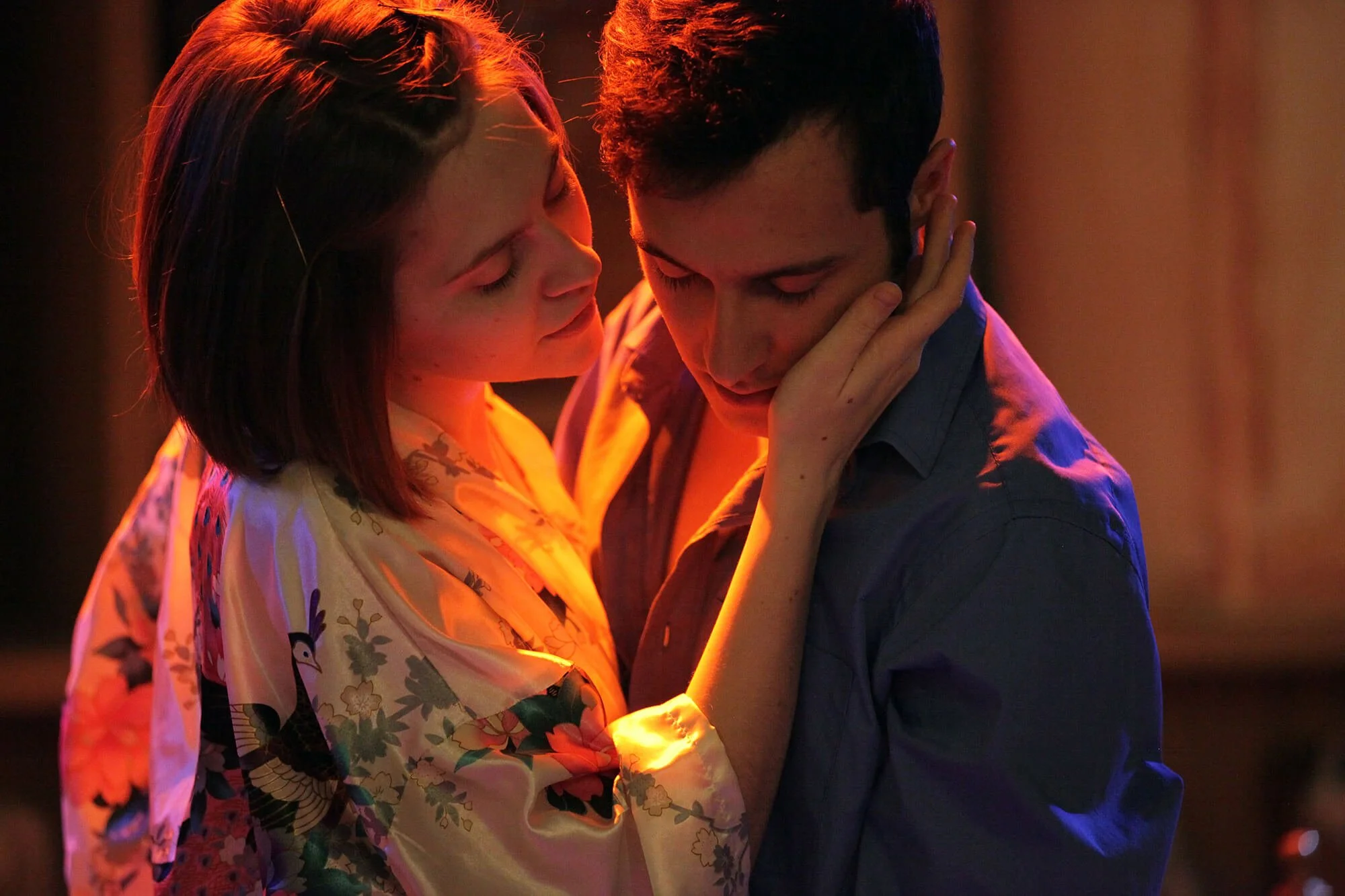 A woman with short dark hair and a floral kimono holds a man's face as they share an intimate moment in a dimly lit setting.