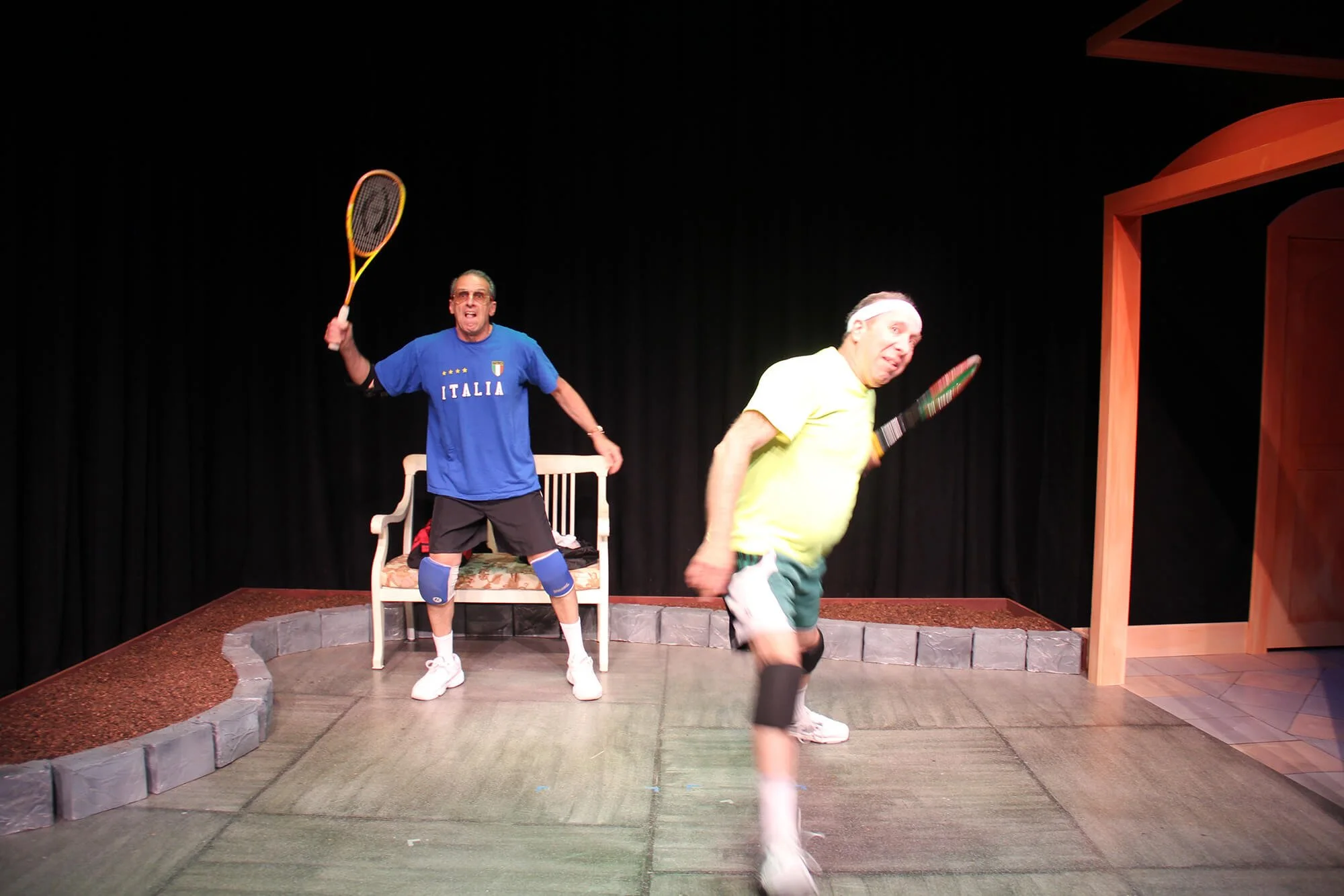 Two men playing indoor tennis with rackets on a stage set. One man in a blue Italy shirt and shorts stands near a bench, holding a tennis racket overhead. The other man, wearing a yellow shirt and shorts, is holding a tennis racket and appears to be 