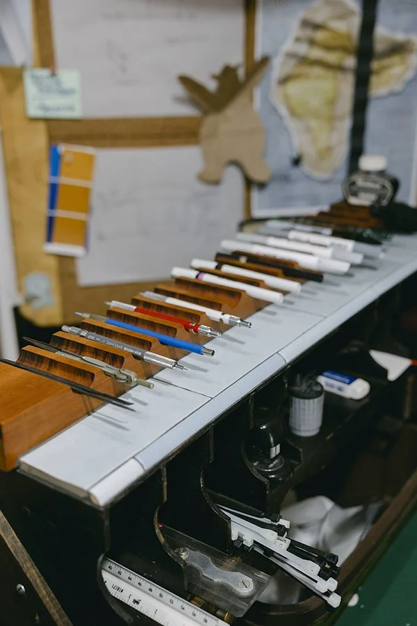 A workbench with drawing instruments, including pens and pencils, lined up on a tilted surface, with a wooden deer decoration and map in the background.