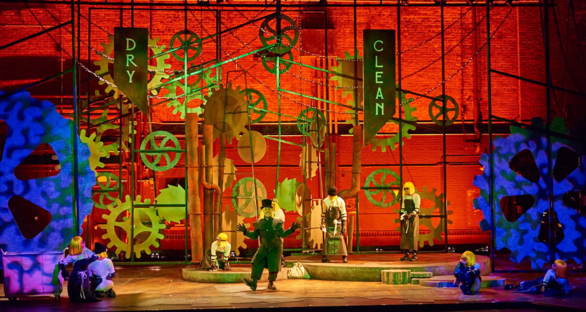 Theater stage with a steampunk-inspired set design featuring large gears, pipes, and industrial elements with a red-orange background. Several performers are in costume, including a person in a black suit and top hat, and children in costumes sitting