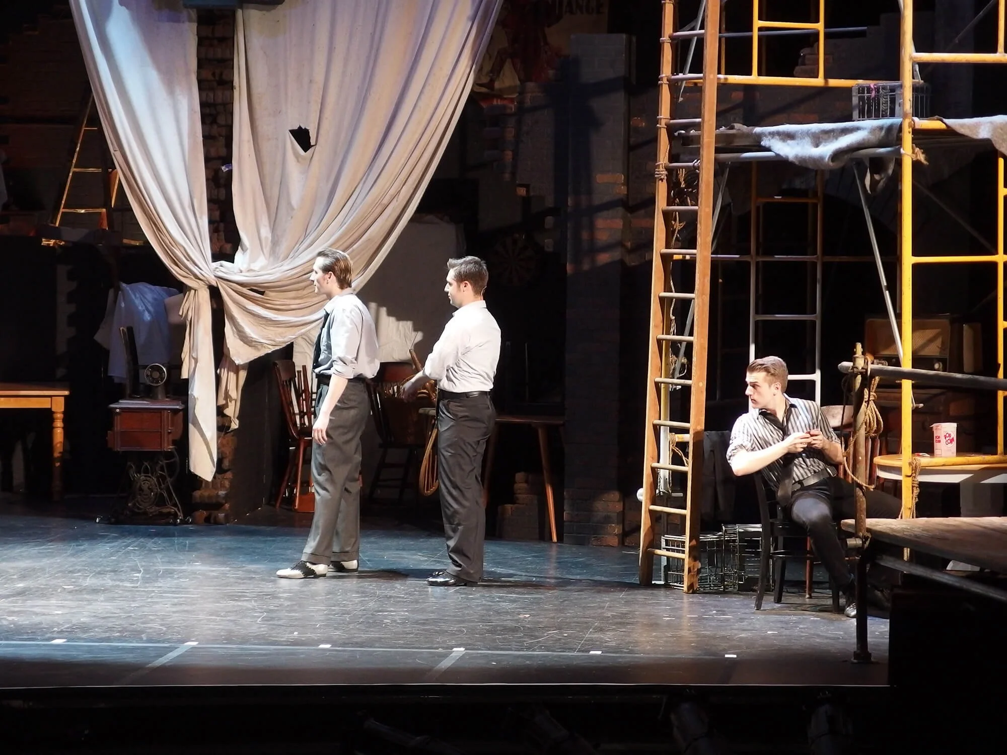 Three men standing on a stage in front of a curtain, with a fourth man sitting to the side near scaffolding and stage equipment.