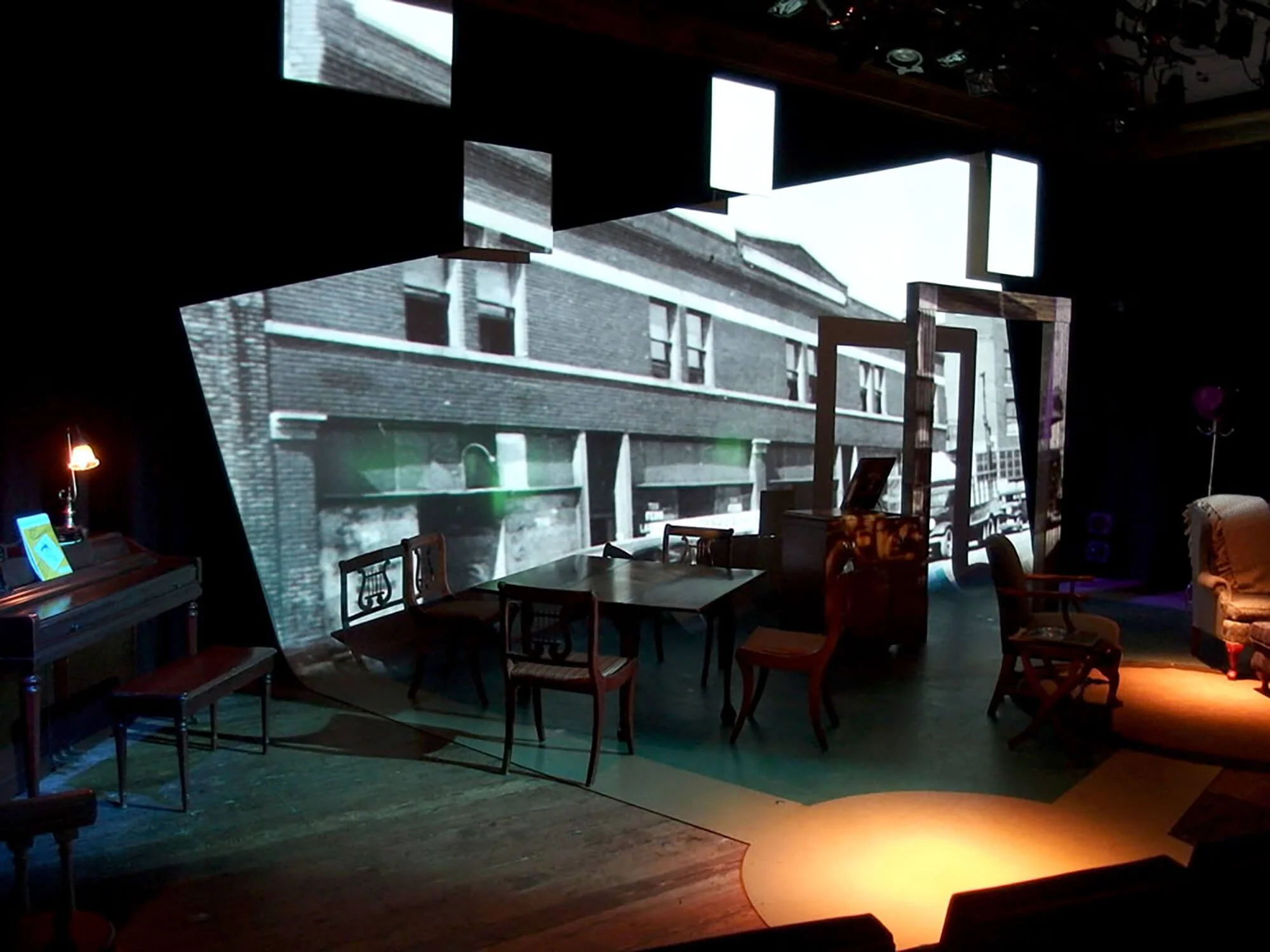 An indoor room with dark lighting, featuring a large black-and-white projected image of a building facade on the wall. The room has antique-style chairs and tables, a piano, and a cozy armchair, creating a vintage ambiance.