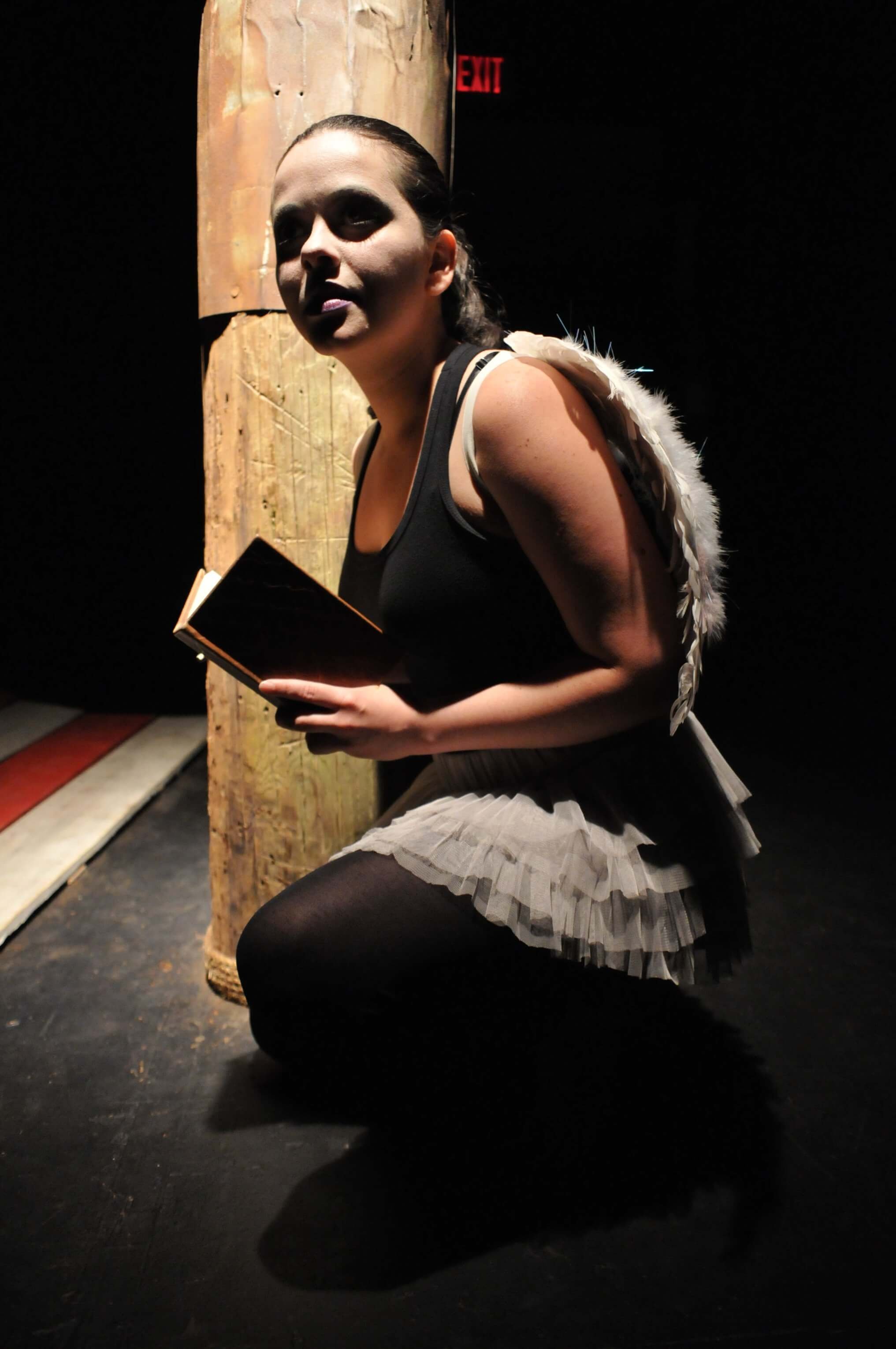 A woman dressed as an angel, wearing a black top and a tutu, kneeling on the floor holding a small book in dim lighting, with a wooden post and an exit sign in the background.