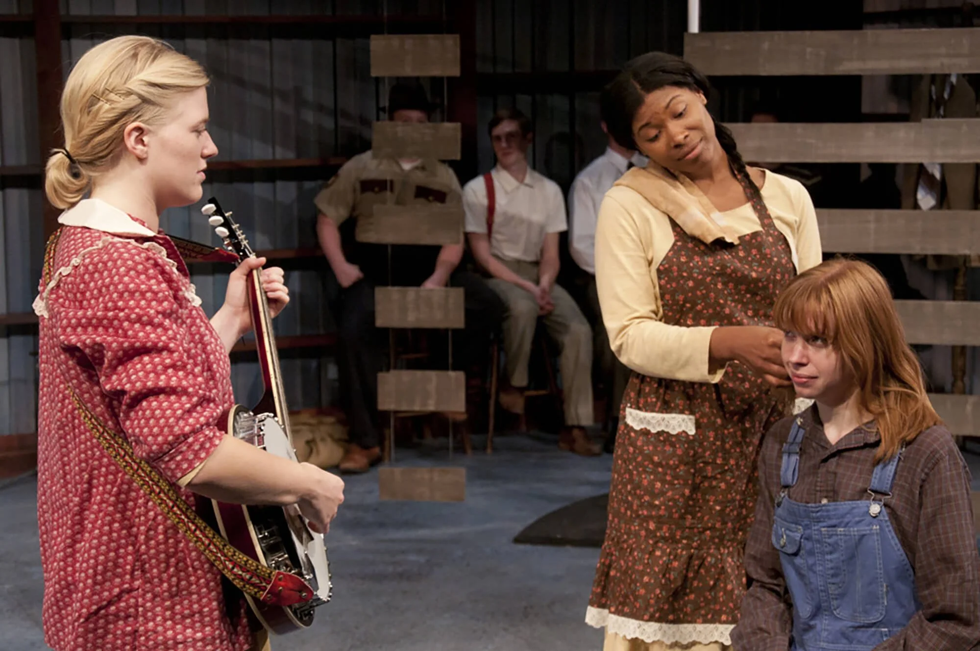 A scene from a play with a young woman holding a banjo, an African American woman speaking to a red-haired girl in denim overalls, and a group of people watching in the background.