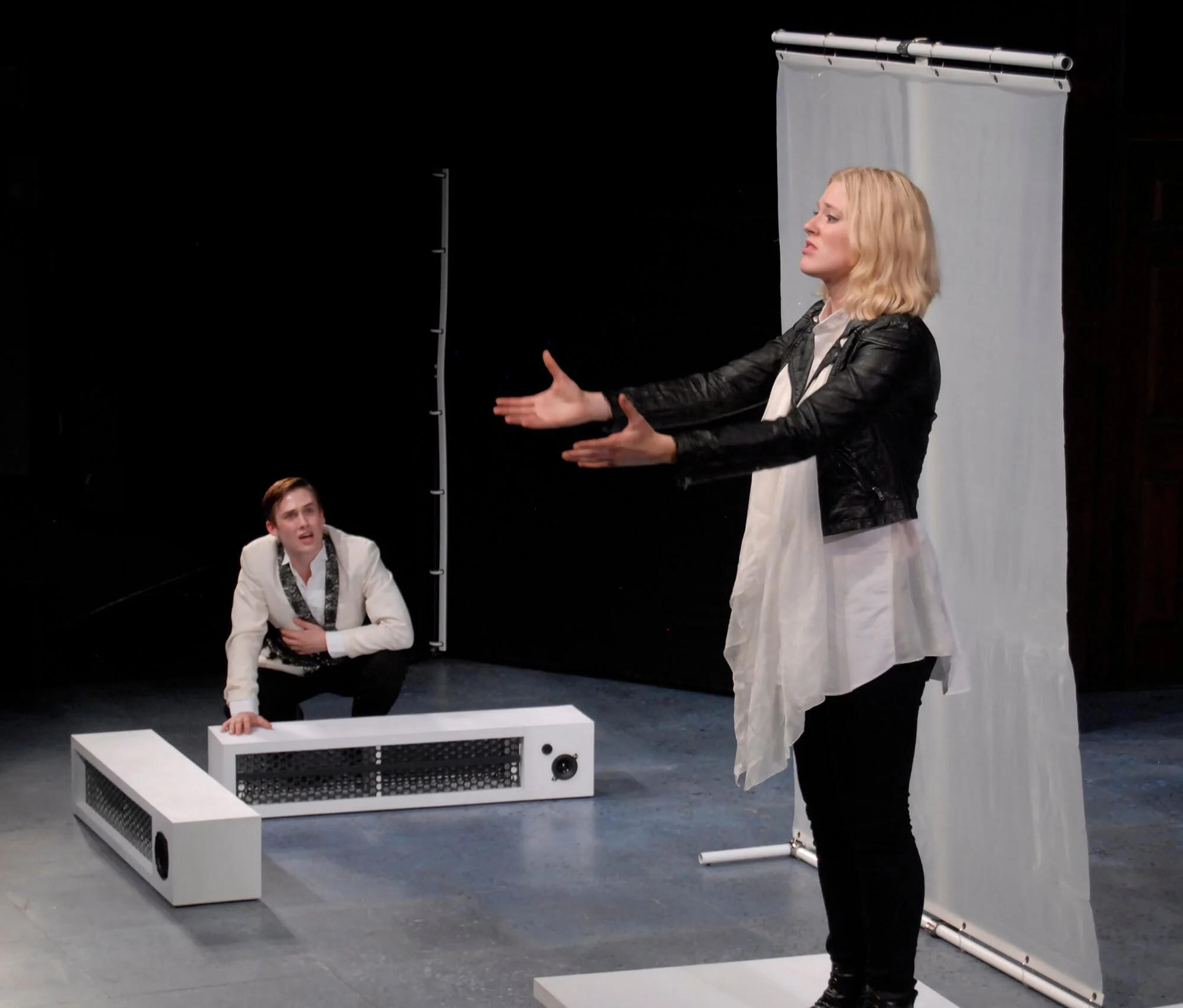 Two women perform on a stage. One woman stands with arms outstretched, and the other woman crouches on the floor, leaning on a white rectangular object. There is a black backdrop and a light gray screen behind them.