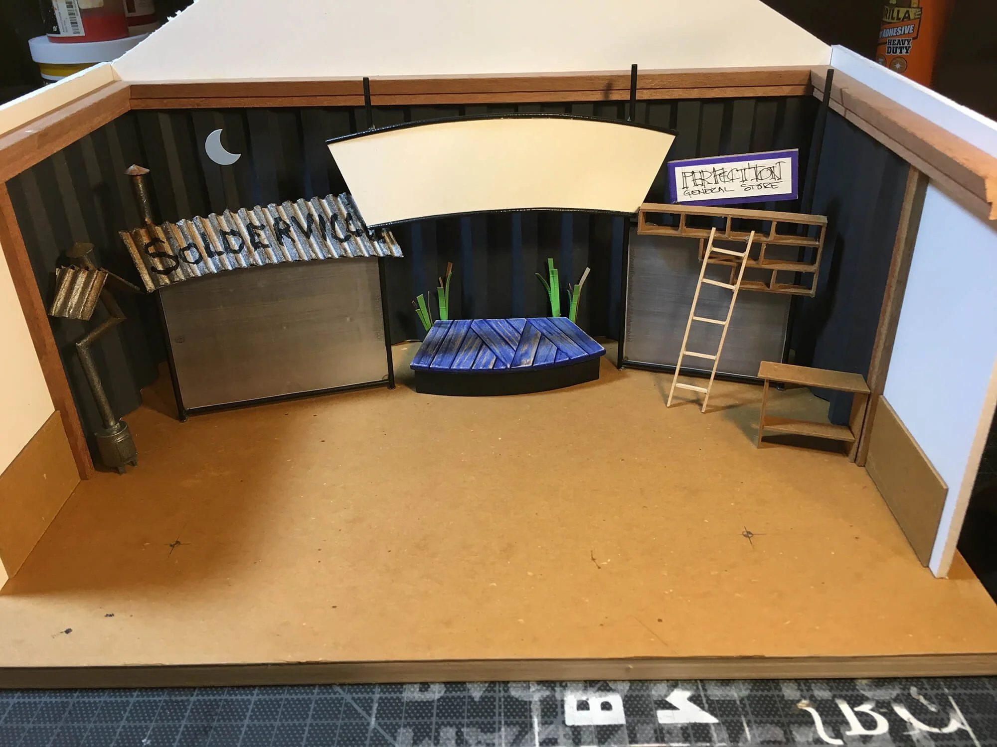 Miniature model of a school science classroom with desks, indoor plants, and signage including a sign that says 'Teacher', a ladder, and a sign reading 'Biology General Store'.