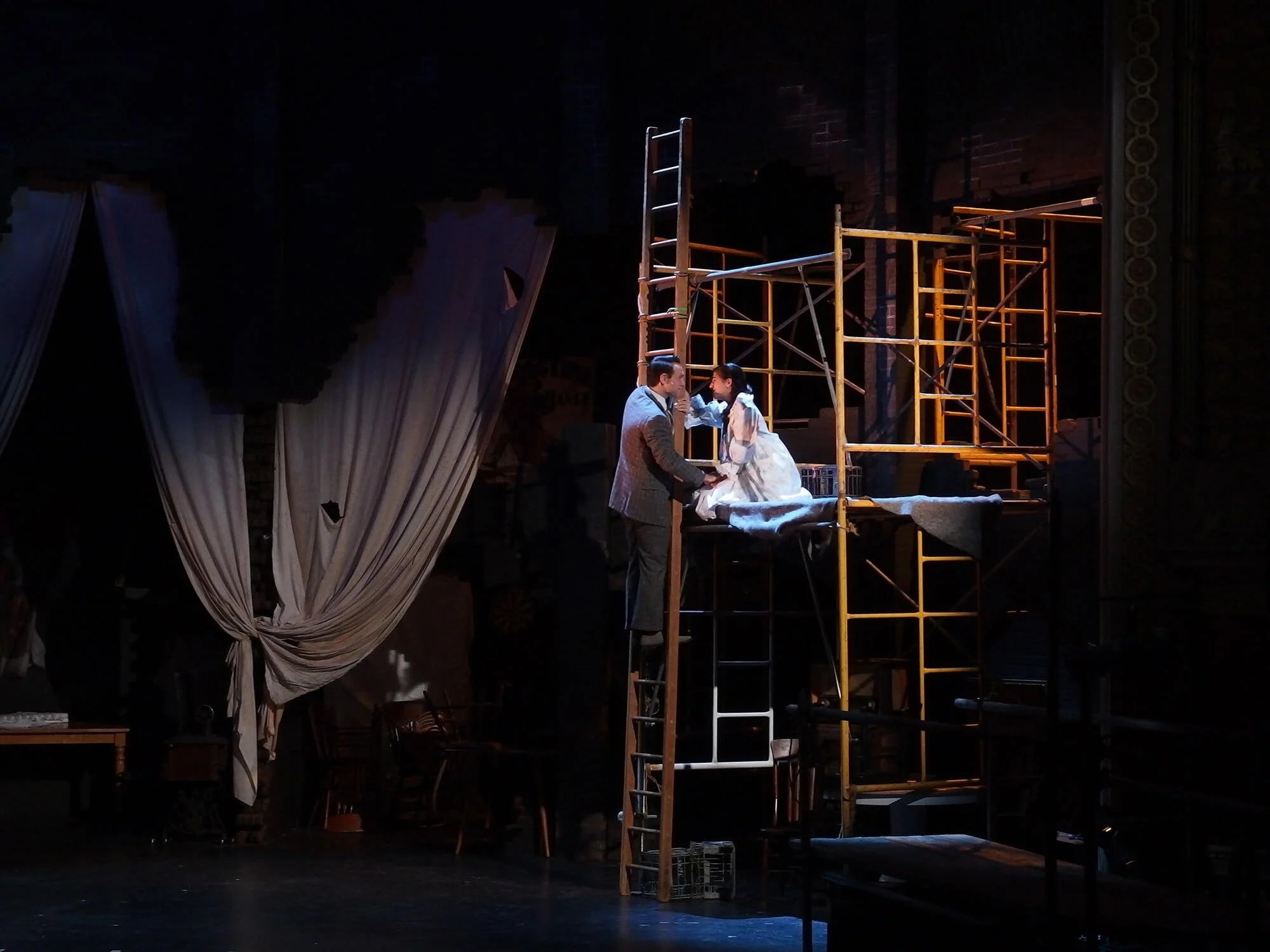 Stage scene with a man and woman on a scaffold, engaging in a conversation as part of a theatrical production, with stage props and curtains in the background.