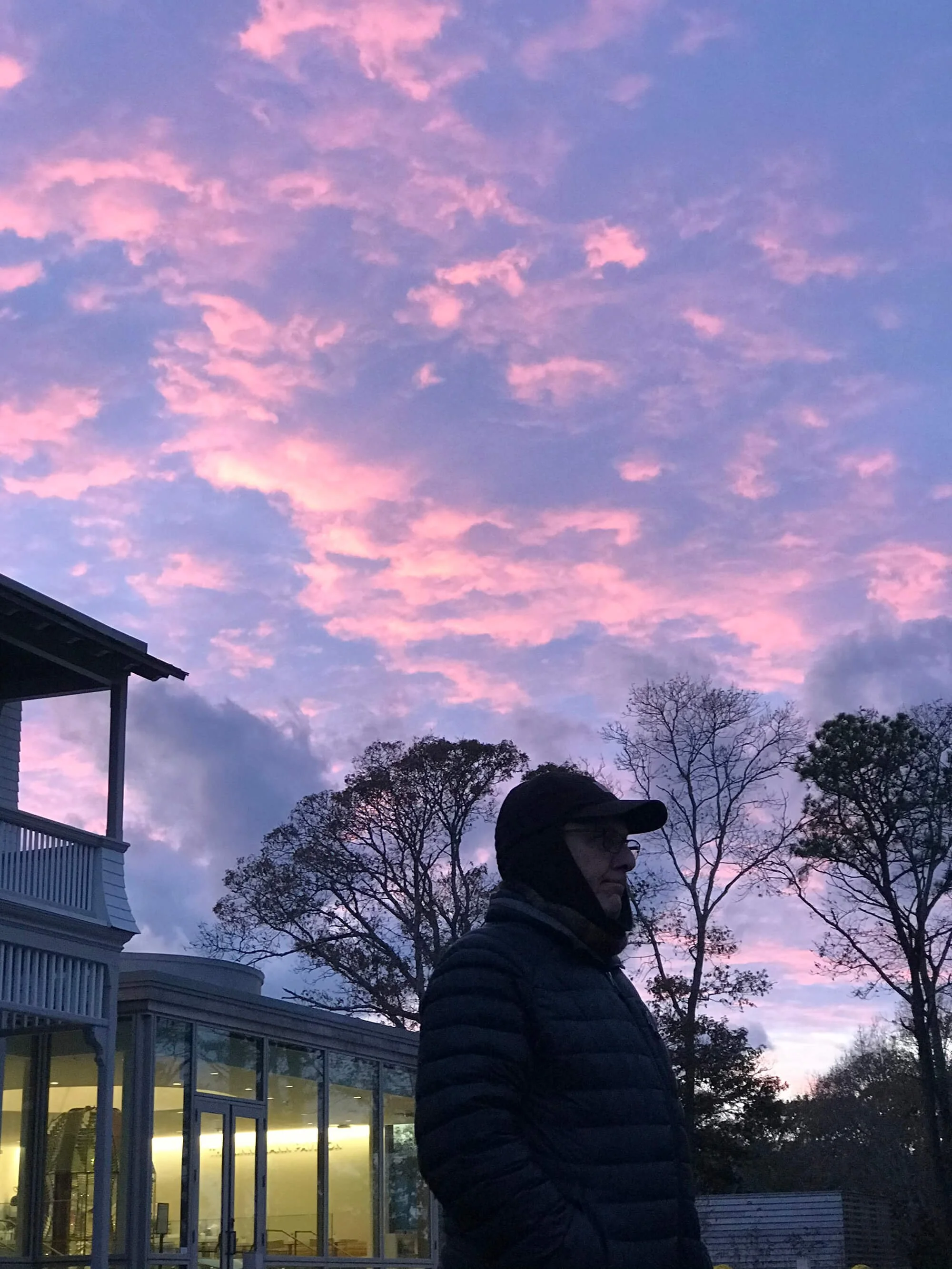 A man wearing a black jacket, cap, and glasses standing outside during sunset with pink and purple clouds, trees, and a building with large windows reflecting light.