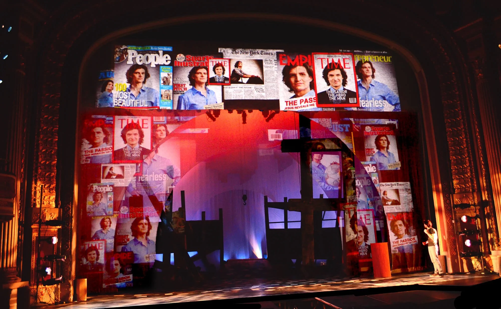 The stage features a large backdrop with magazines and newspaper covers, including TIME and People, displaying a woman with dark hair. The stage is set with a silhouette of a boat and dock, illuminated with red and blue lights, within an ornate theat