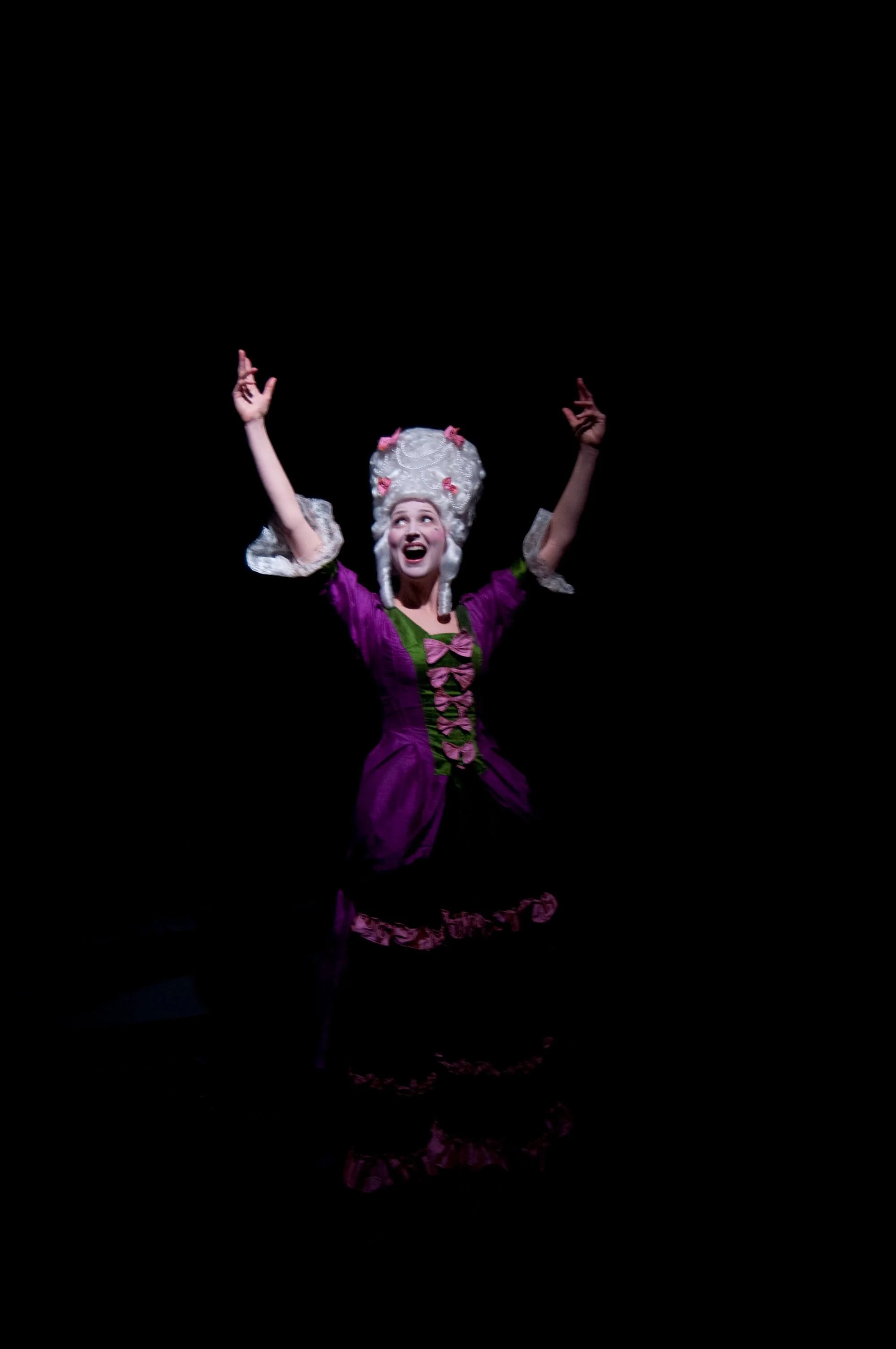 A woman dressed in a colorful, historical costume with a purple and green dress and elaborate white wig, performing on a dark stage with arms raised and an excited expression.