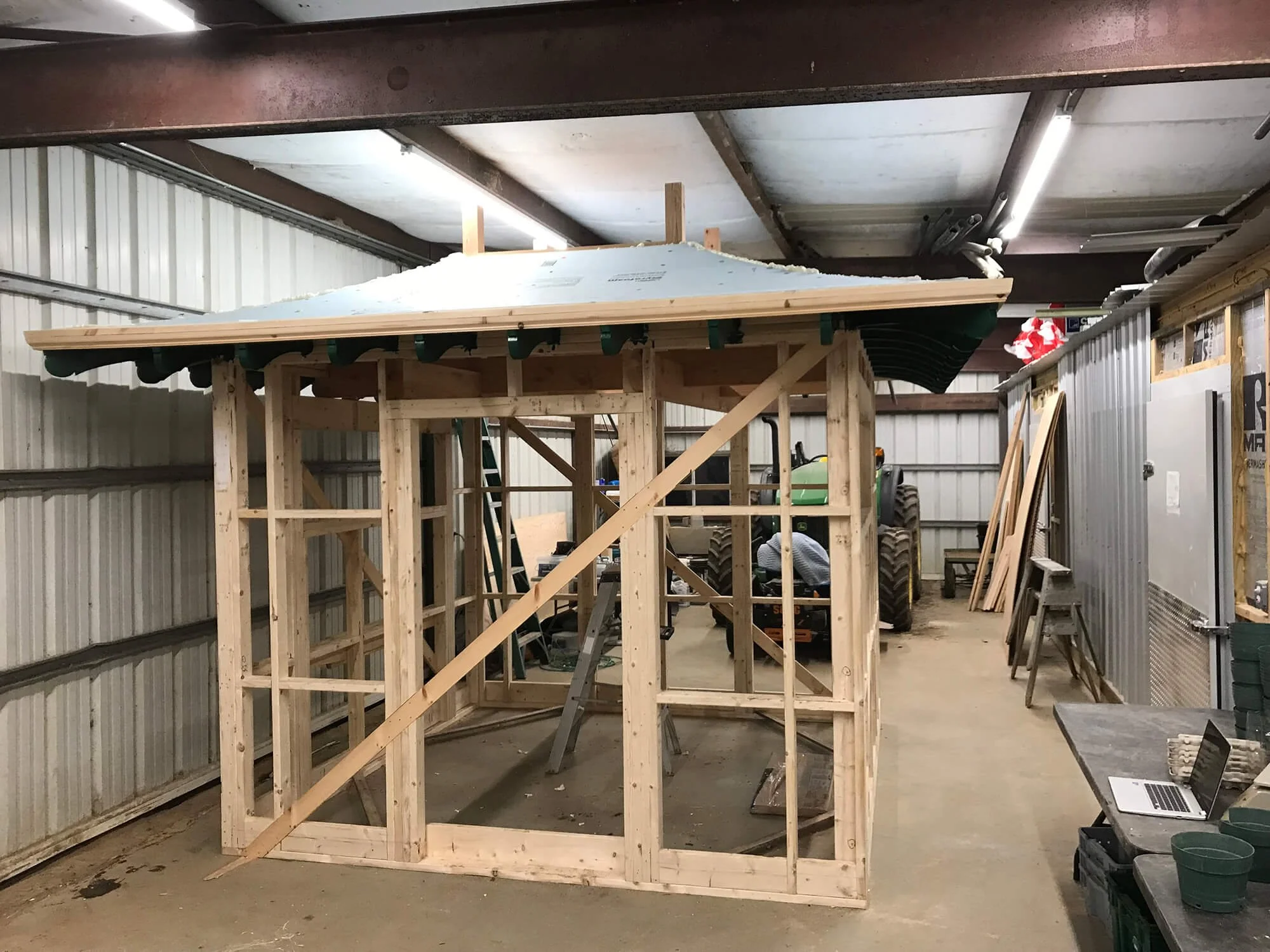 Wooden framework of a small building or shed under construction inside a workshop or garage with tools and equipment visible.